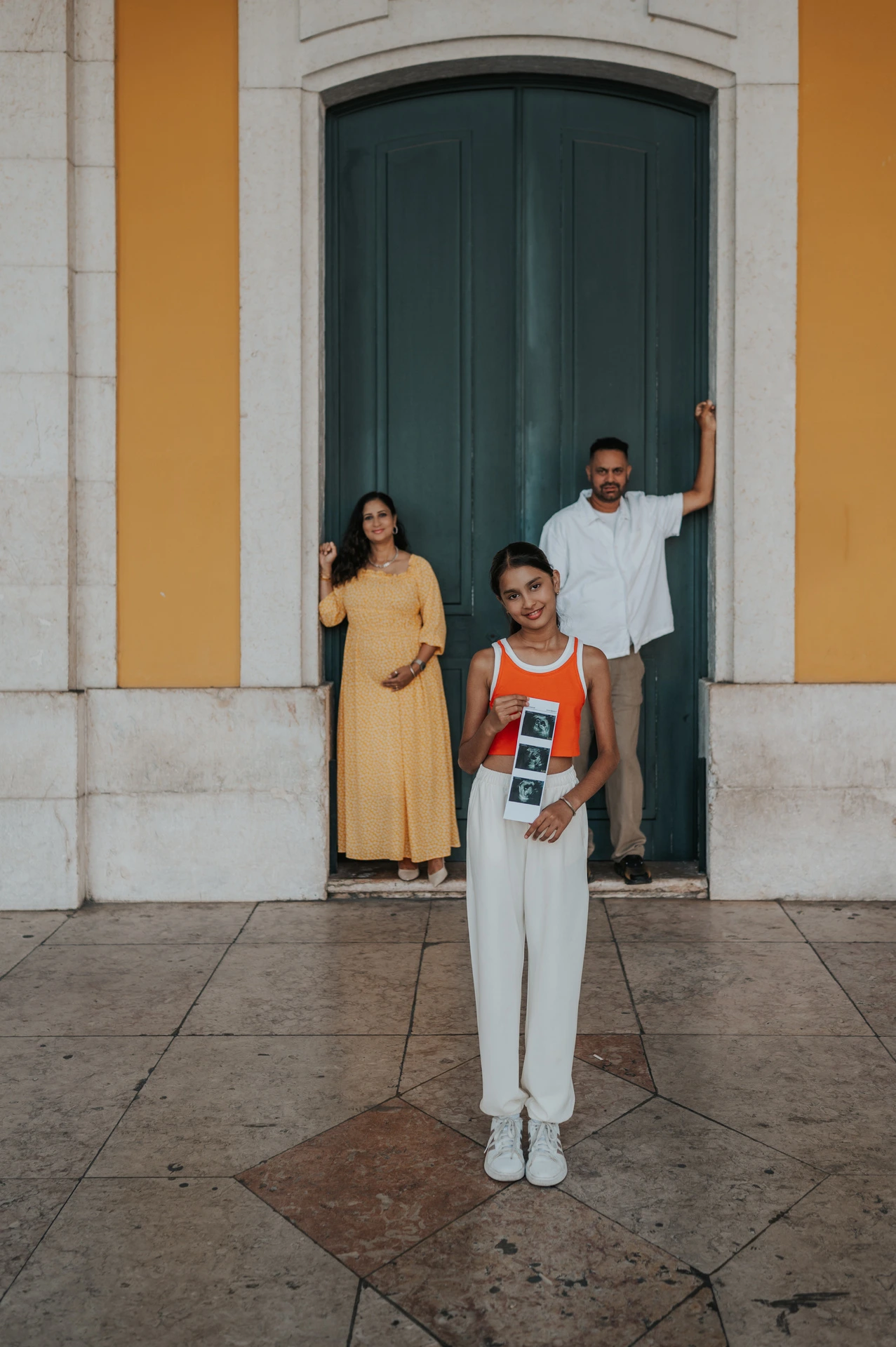 The daughter holding ultrasound photographs with her parents in the doorway behind her during their family and maternity photoshoot in Lisbon.
