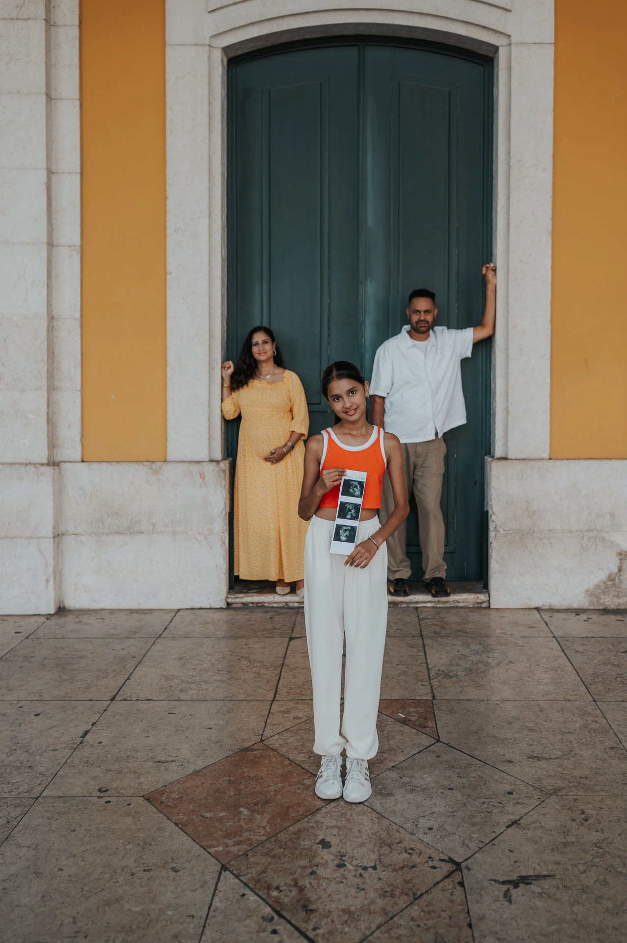 The daughter holding ultrasound photographs with her parents in the doorway behind her during their family and maternity photoshoot in Lisbon.