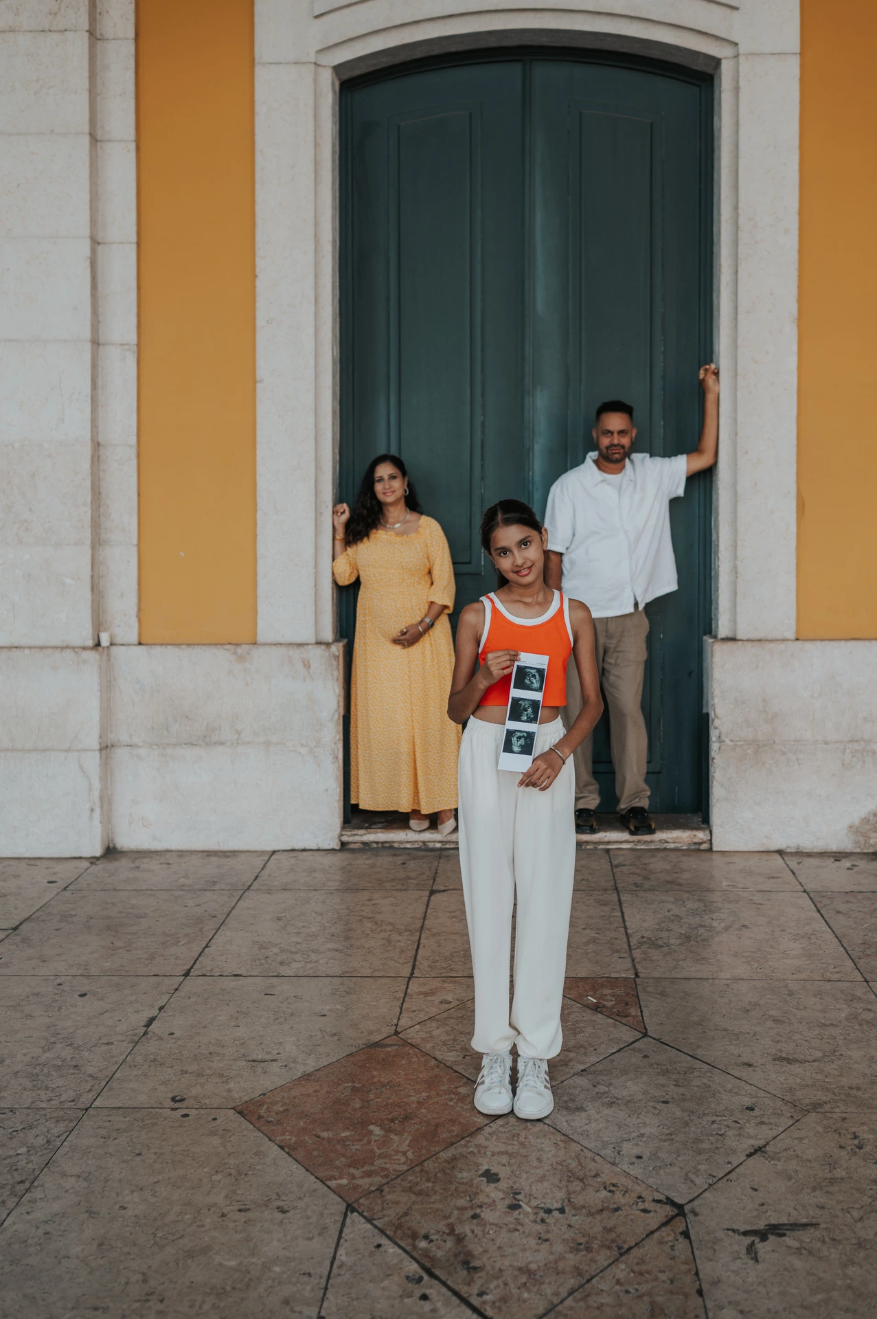 The daughter holding ultrasound photographs with her parents in the doorway behind her during their family and maternity photoshoot in Lisbon.