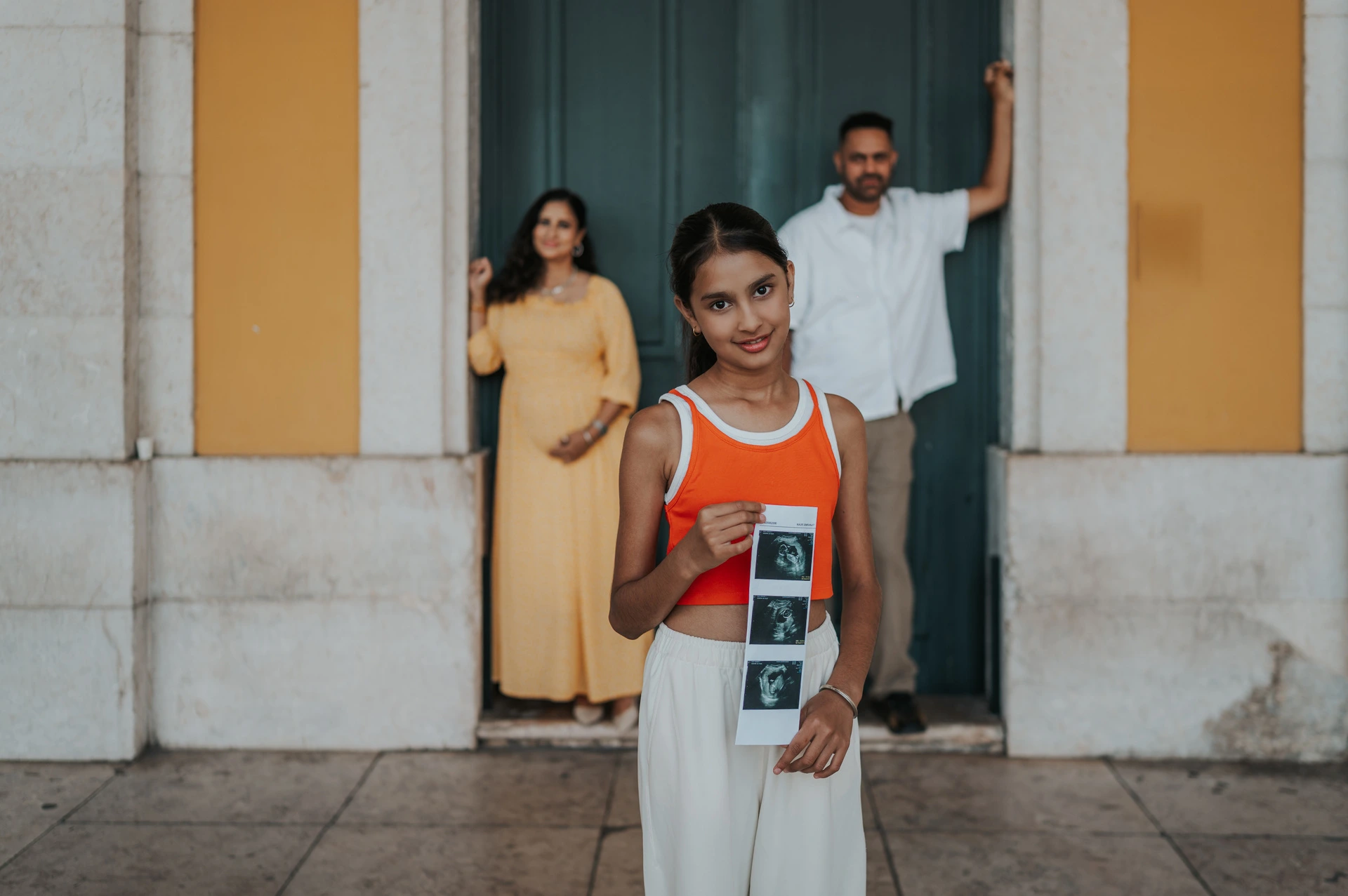 The daughter holding ultrasound photographs with her parents in the doorway behind her during their family and maternity photoshoot in Lisbon.