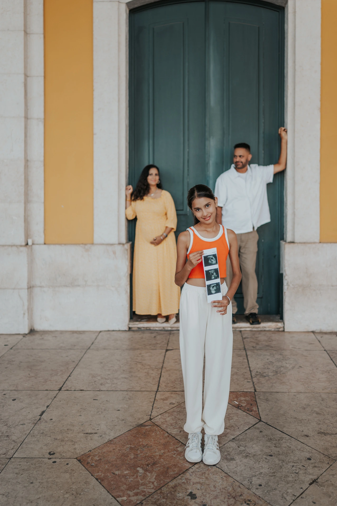 The daughter holding ultrasound photographs with her parents in the doorway behind her during their family and maternity photoshoot in Lisbon.