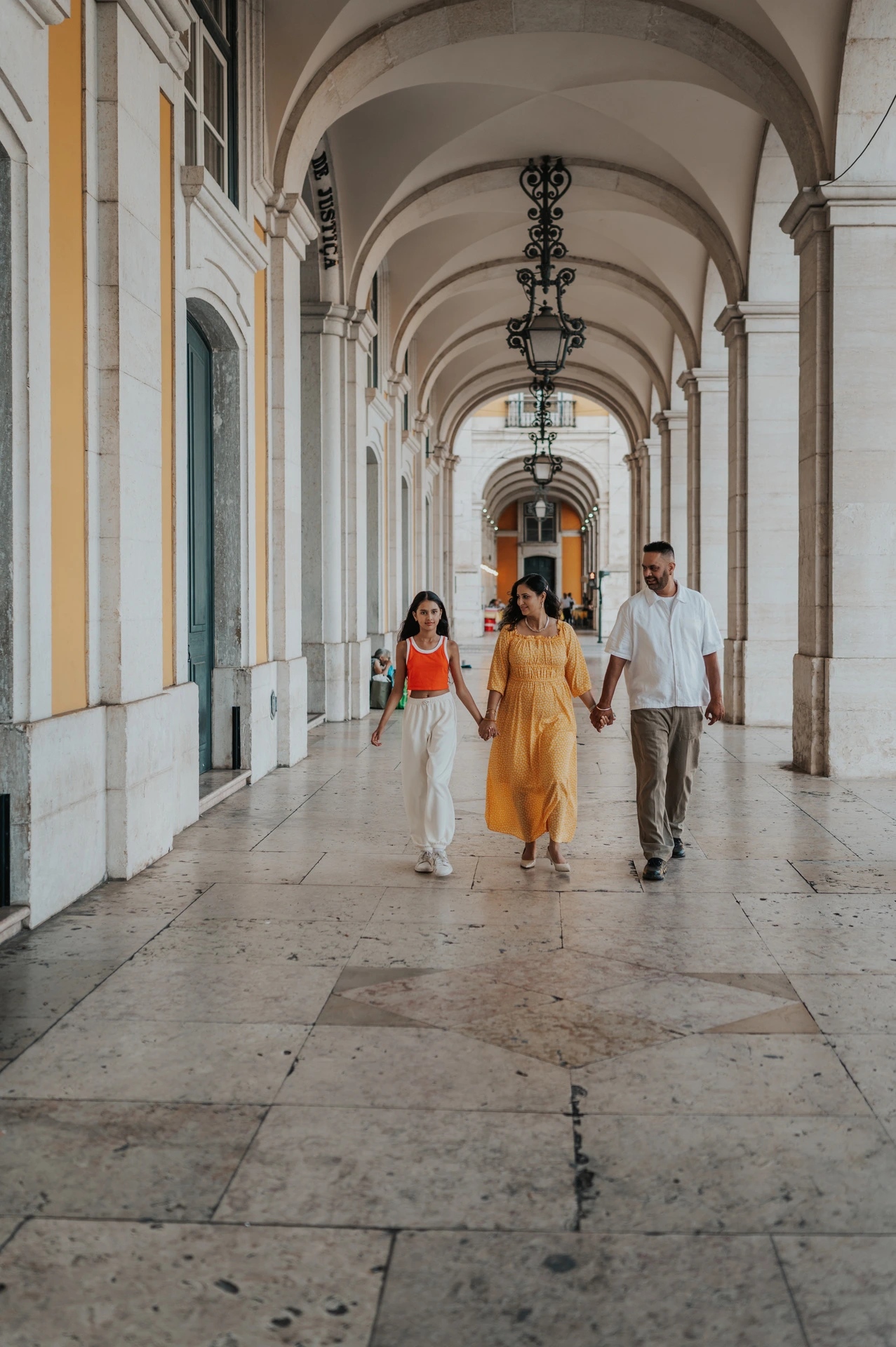 The parents and daughter walking hand in hand beneath the stone arcade during their family and maternity photoshoot in Lisbon.
