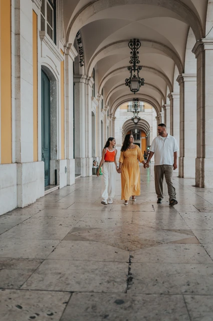 A family walk beneath the arcade | Lisbon family and maternity photoshoot The parents and daughter walking hand in hand beneath the stone arcade during their family and maternity photoshoot in Lisbon.