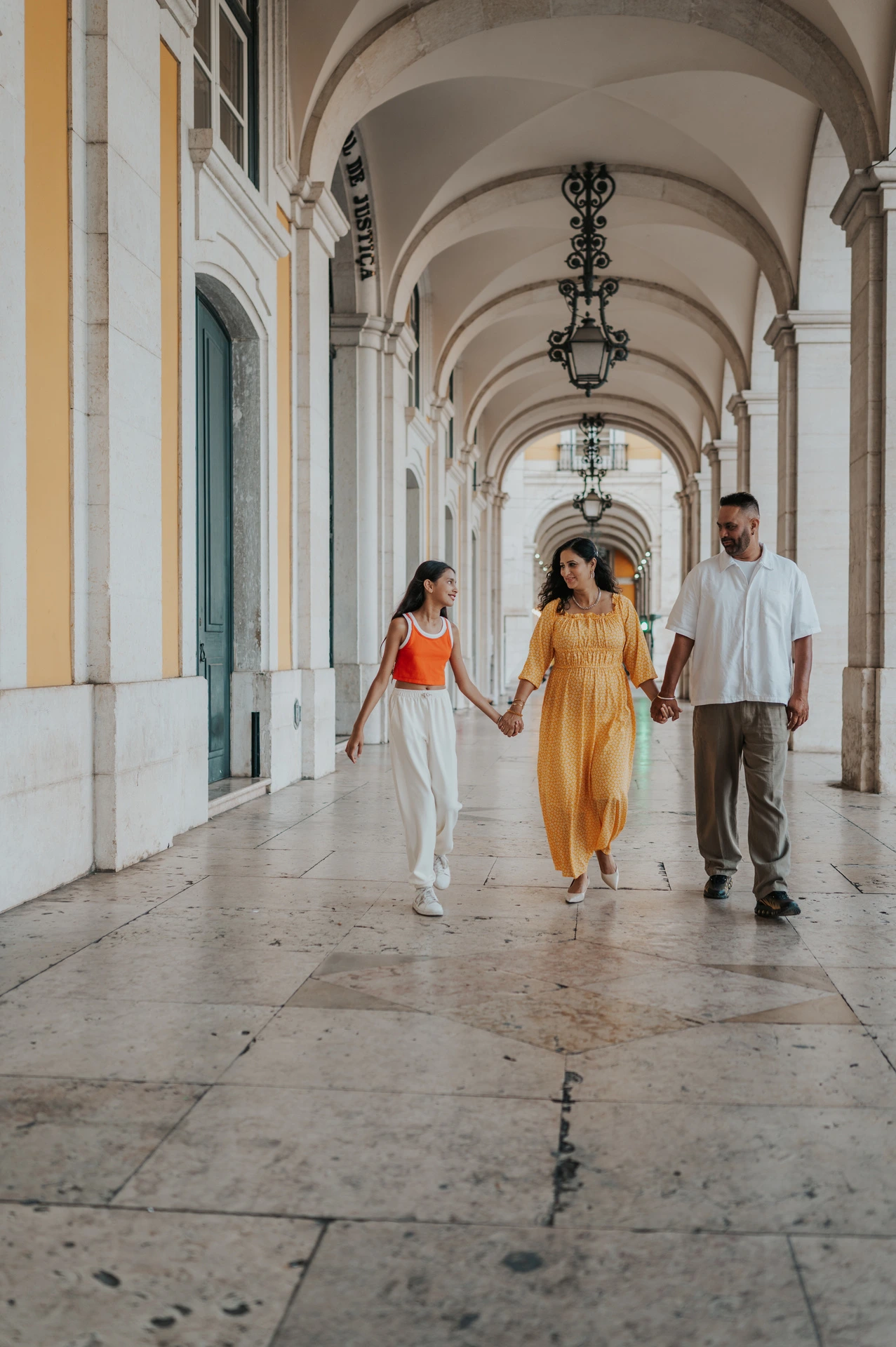 The parents and daughter walking hand in hand beneath the stone arcade during their family and maternity photoshoot in Lisbon.