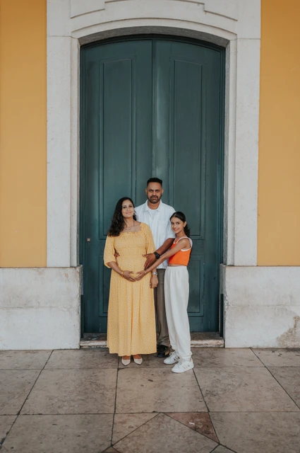 A family portrait at the green door | Lisbon family and maternity photoshoot The parents and their daughter standing close together beside the green door at Praça do Comércio during their family and maternity photoshoot in Lisbon.