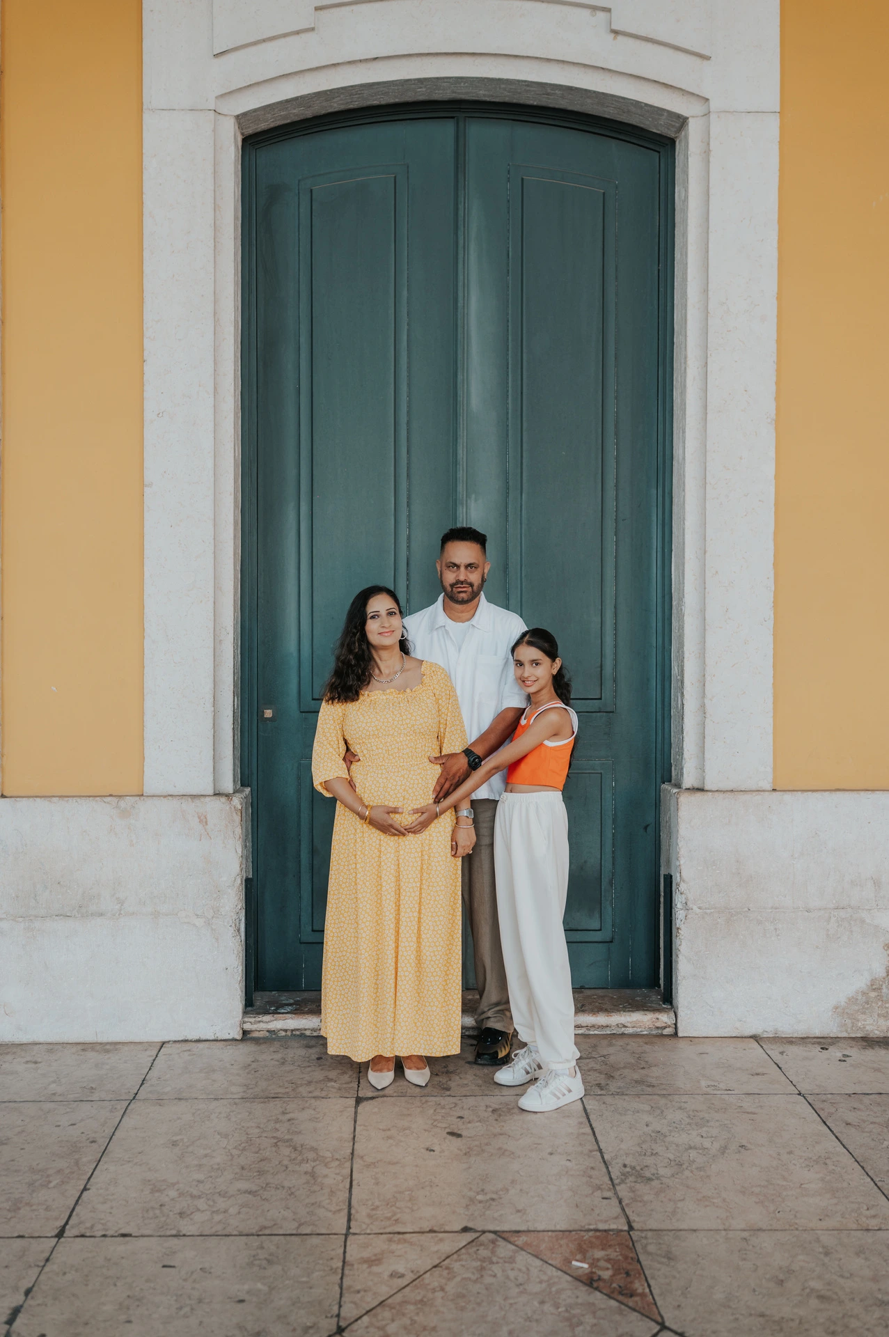 The parents and their daughter standing close together beside the green door at Praça do Comércio during their family and maternity photoshoot in Lisbon.