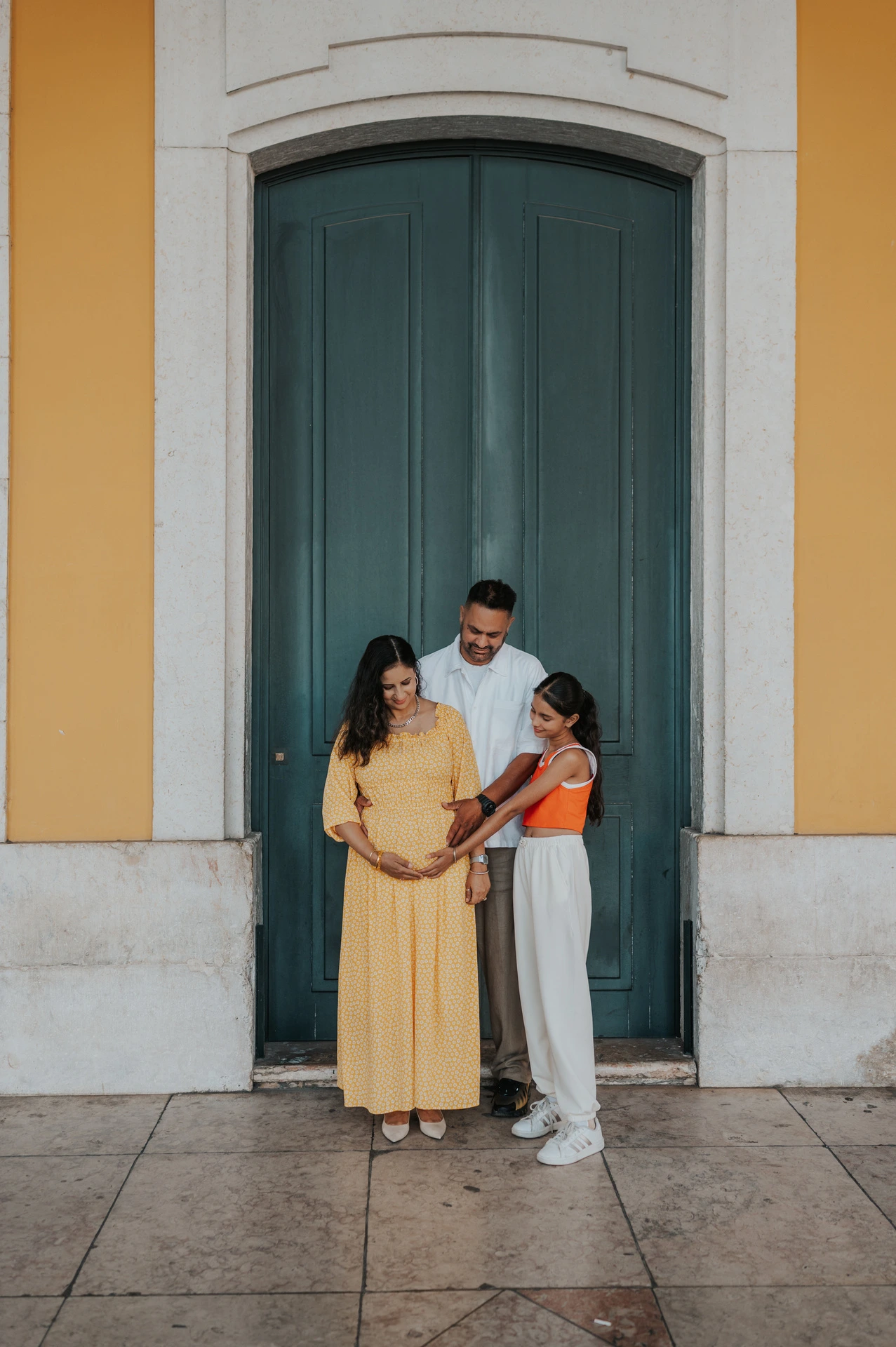 The parents and their daughter standing close together beside the green door at Praça do Comércio during their family and maternity photoshoot in Lisbon.