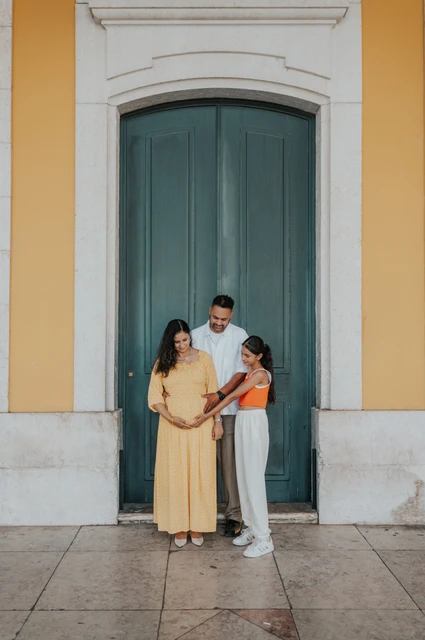 A family portrait at the green door | Lisbon family and maternity photoshoot The parents and their daughter standing close together beside the green door at Praça do Comércio during their family and maternity photoshoot in Lisbon.