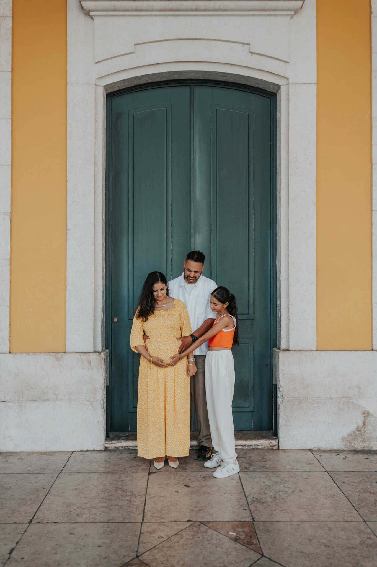 The parents and their daughter standing close together beside the green door at Praça do Comércio during their family and maternity photoshoot in Lisbon.