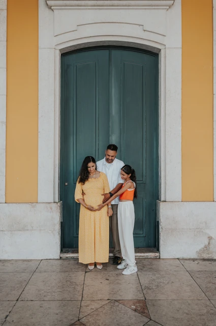 A family portrait at the green door | Lisbon family and maternity photoshoot The parents and their daughter standing close together beside the green door at Praça do Comércio during their family and maternity photoshoot in Lisbon.