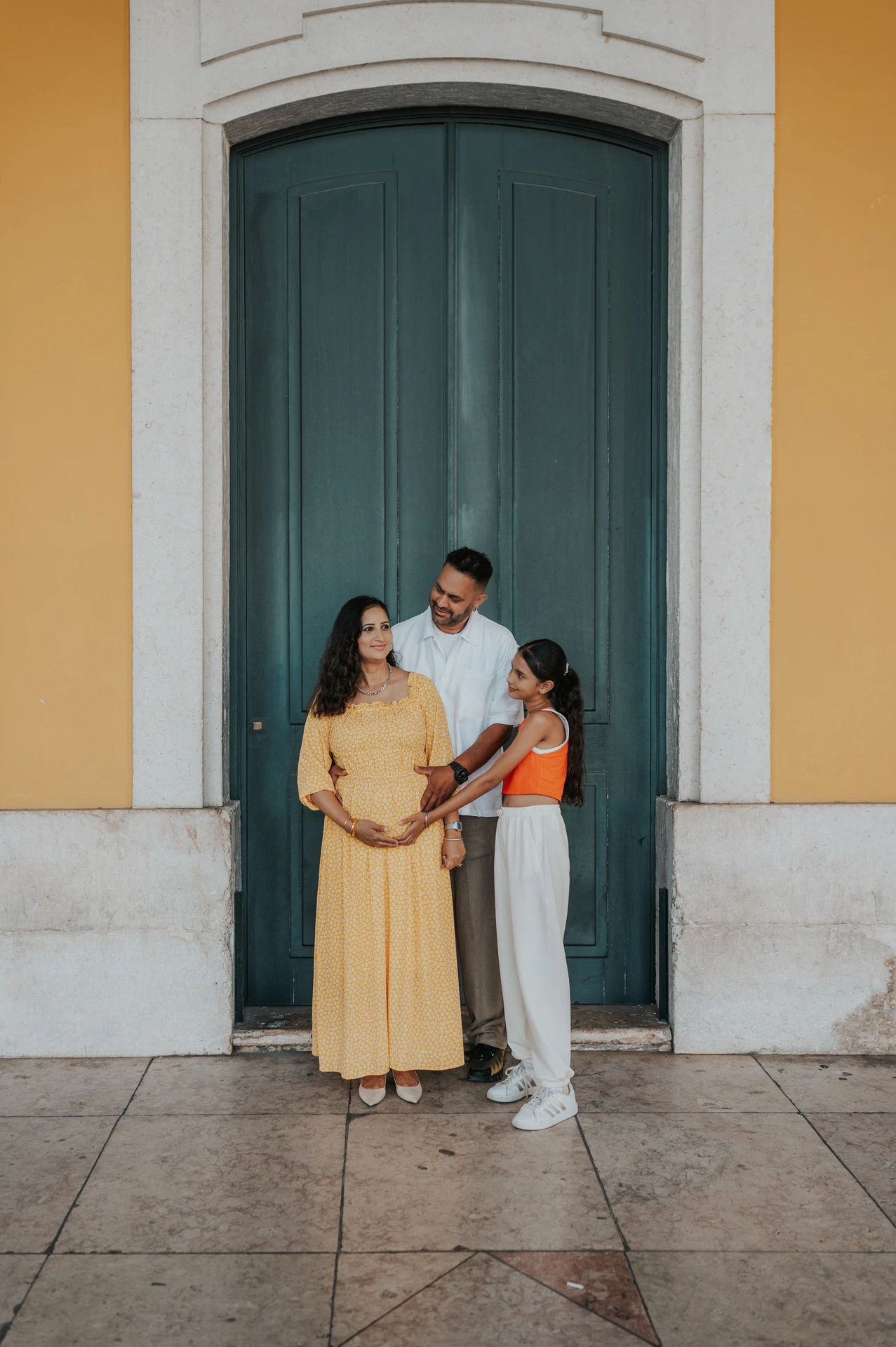The parents and their daughter standing close together beside the green door at Praça do Comércio during their family and maternity photoshoot in Lisbon.