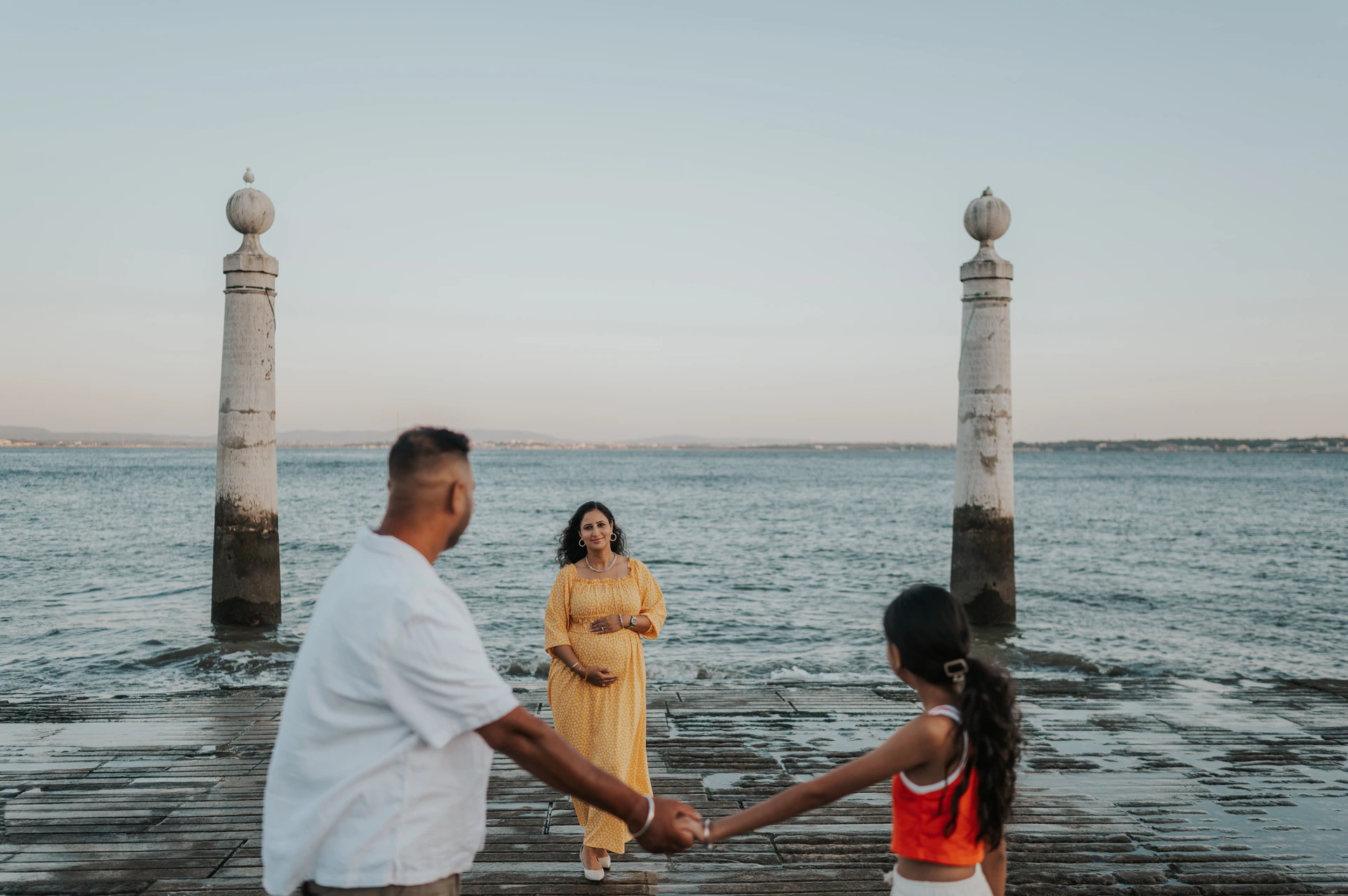 The father and daughter holding hands with the expectant mother beside the river during their family and maternity photoshoot in Lisbon.