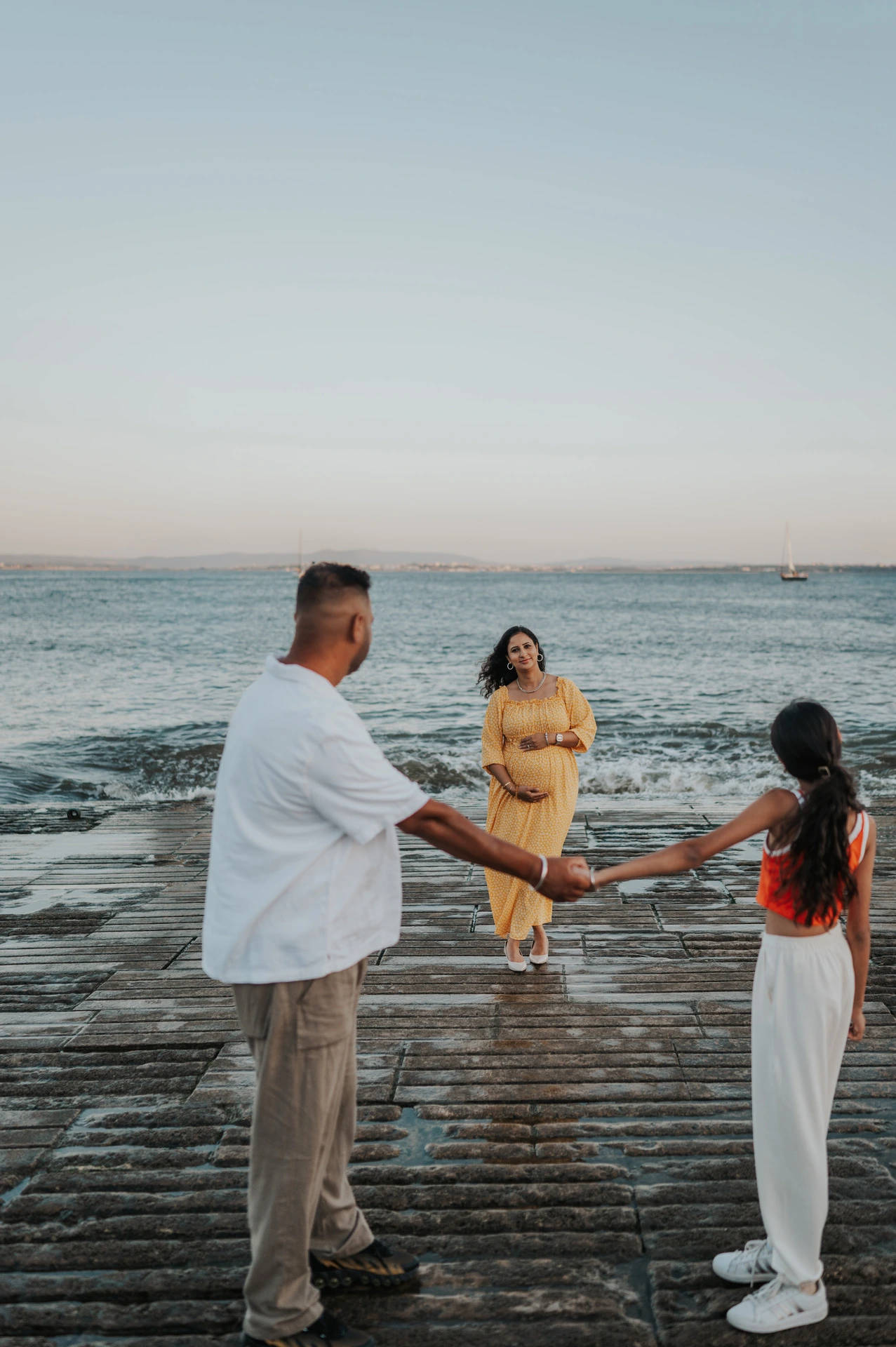The father and daughter holding hands with the expectant mother beside the river during their family and maternity photoshoot in Lisbon.