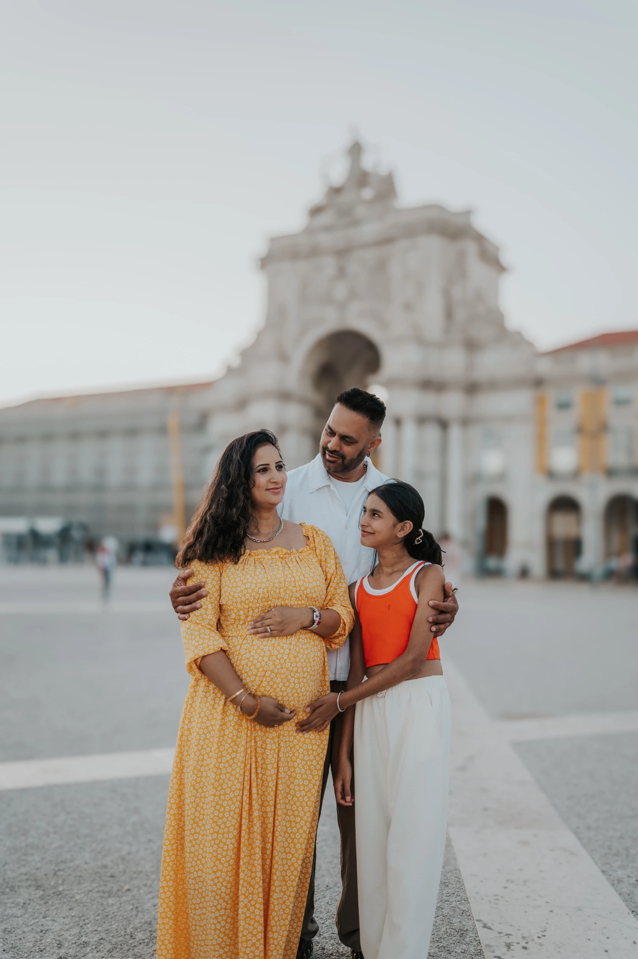 The family standing close together and smiling in front of Praça do Comércio’s arch during their family and maternity photoshoot in Lisbon.