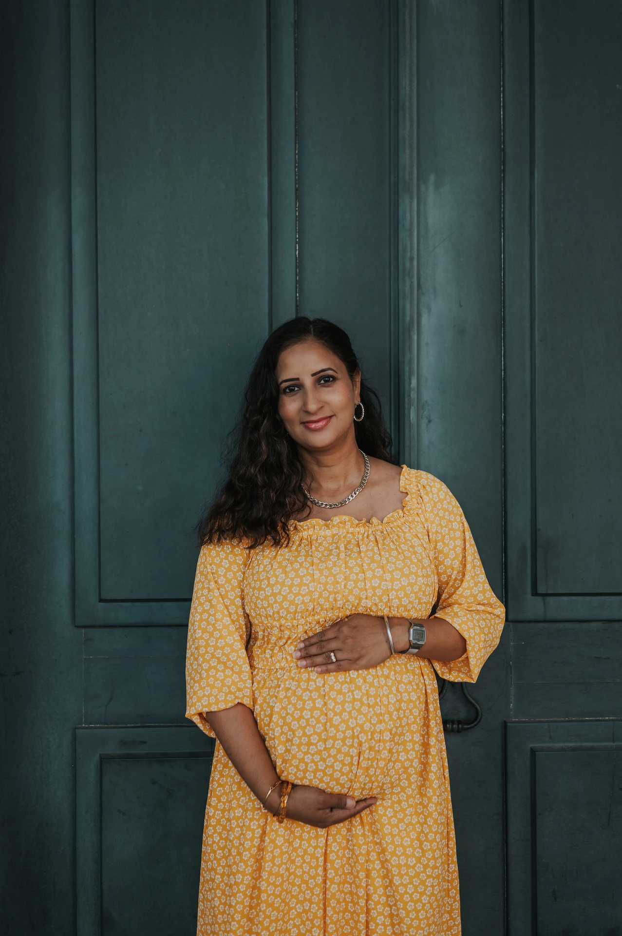 The expectant mother holding her belly in front of the tall green door at Praça do Comércio during their family and maternity photoshoot in Lisbon.