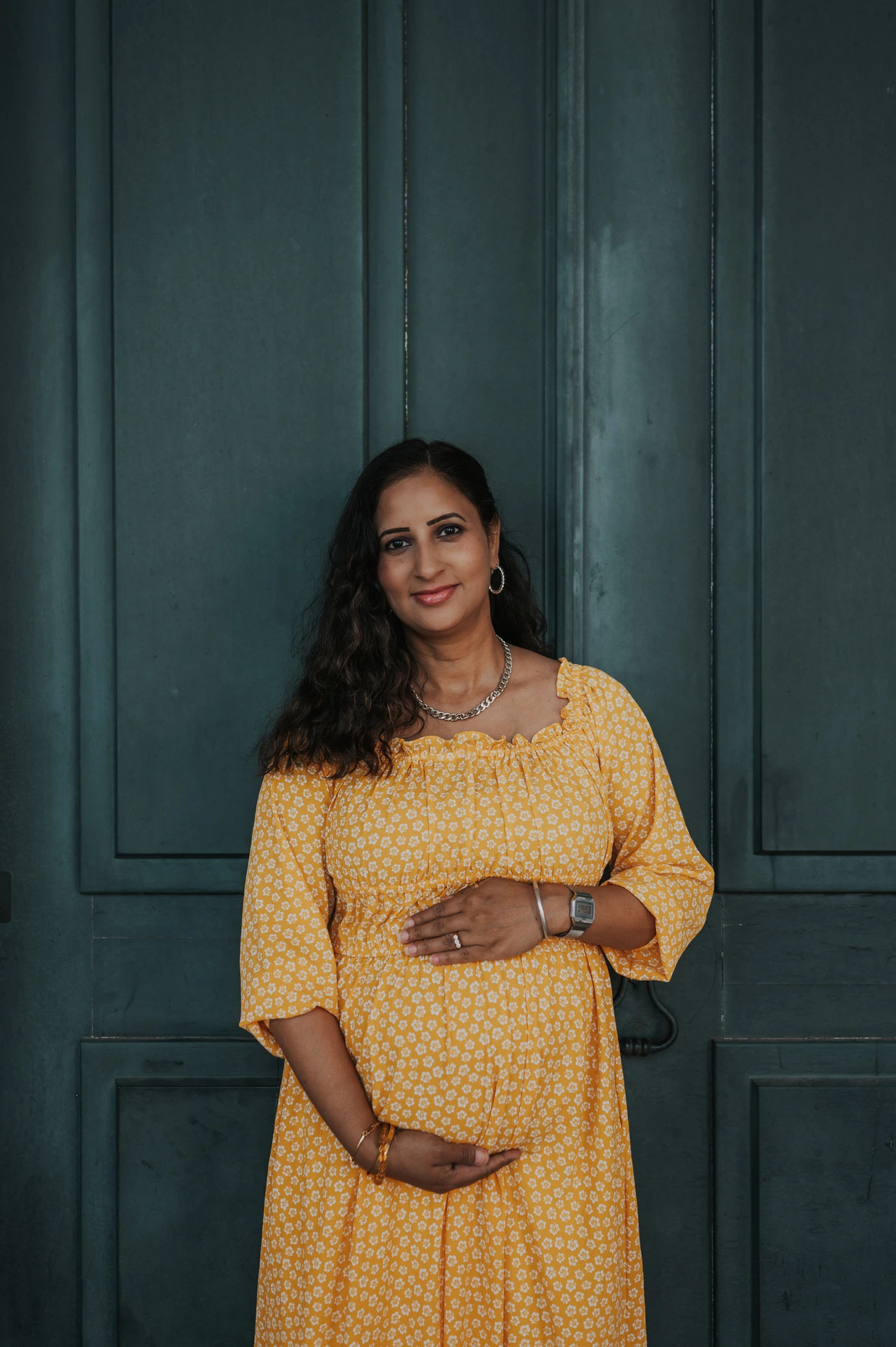The expectant mother holding her belly in front of the tall green door at Praça do Comércio during their family and maternity photoshoot in Lisbon.