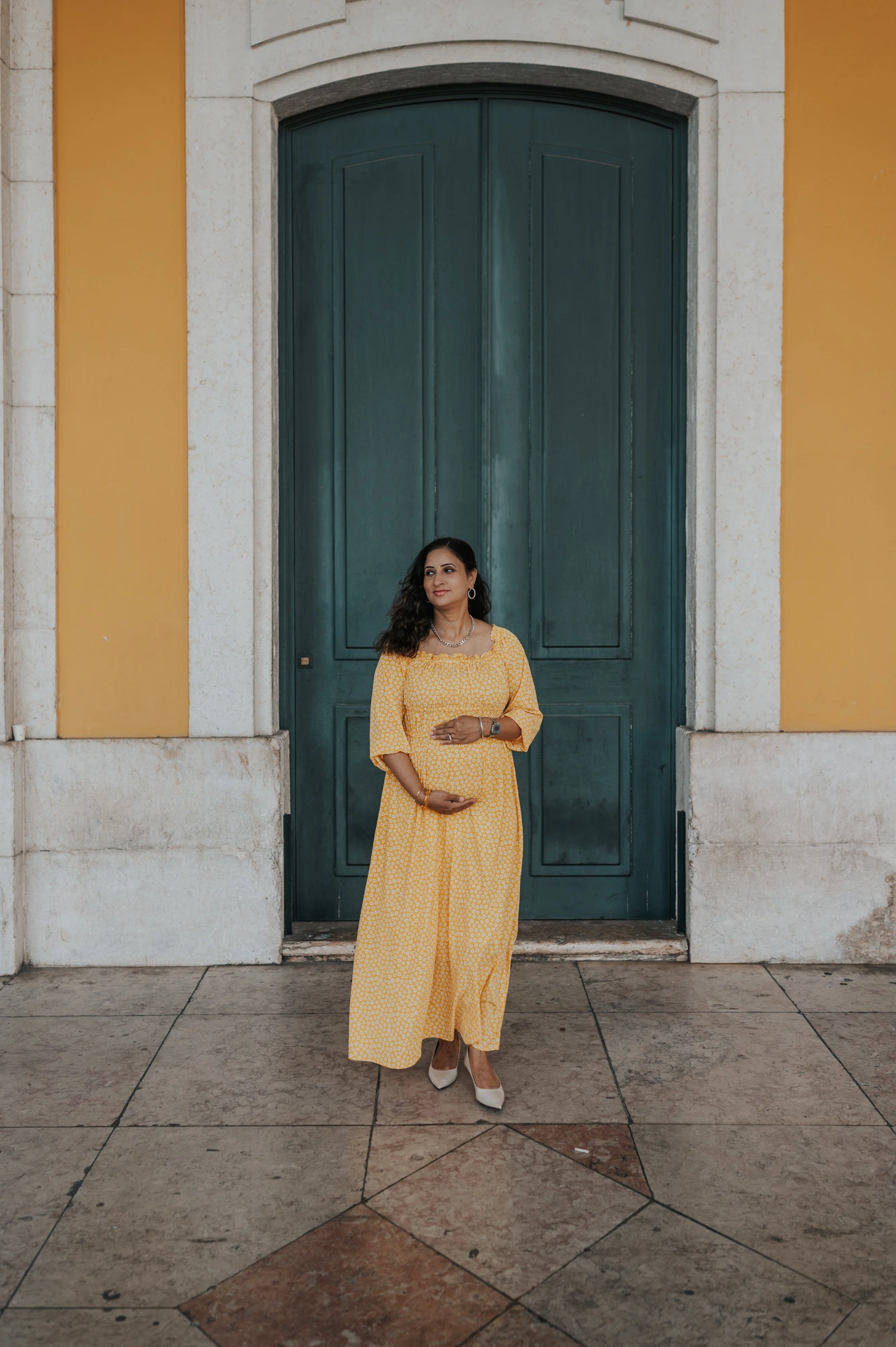The expectant mother holding her belly in front of the tall green door at Praça do Comércio during their family and maternity photoshoot in Lisbon.