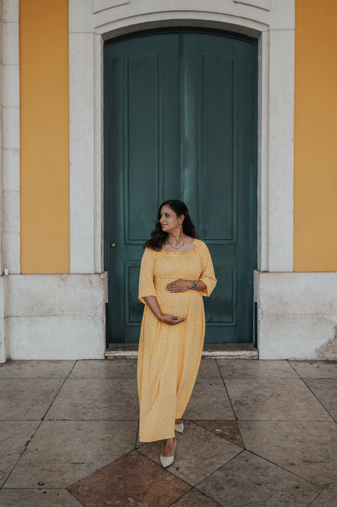 The expectant mother holding her belly in front of the tall green door at Praça do Comércio during their family and maternity photoshoot in Lisbon.