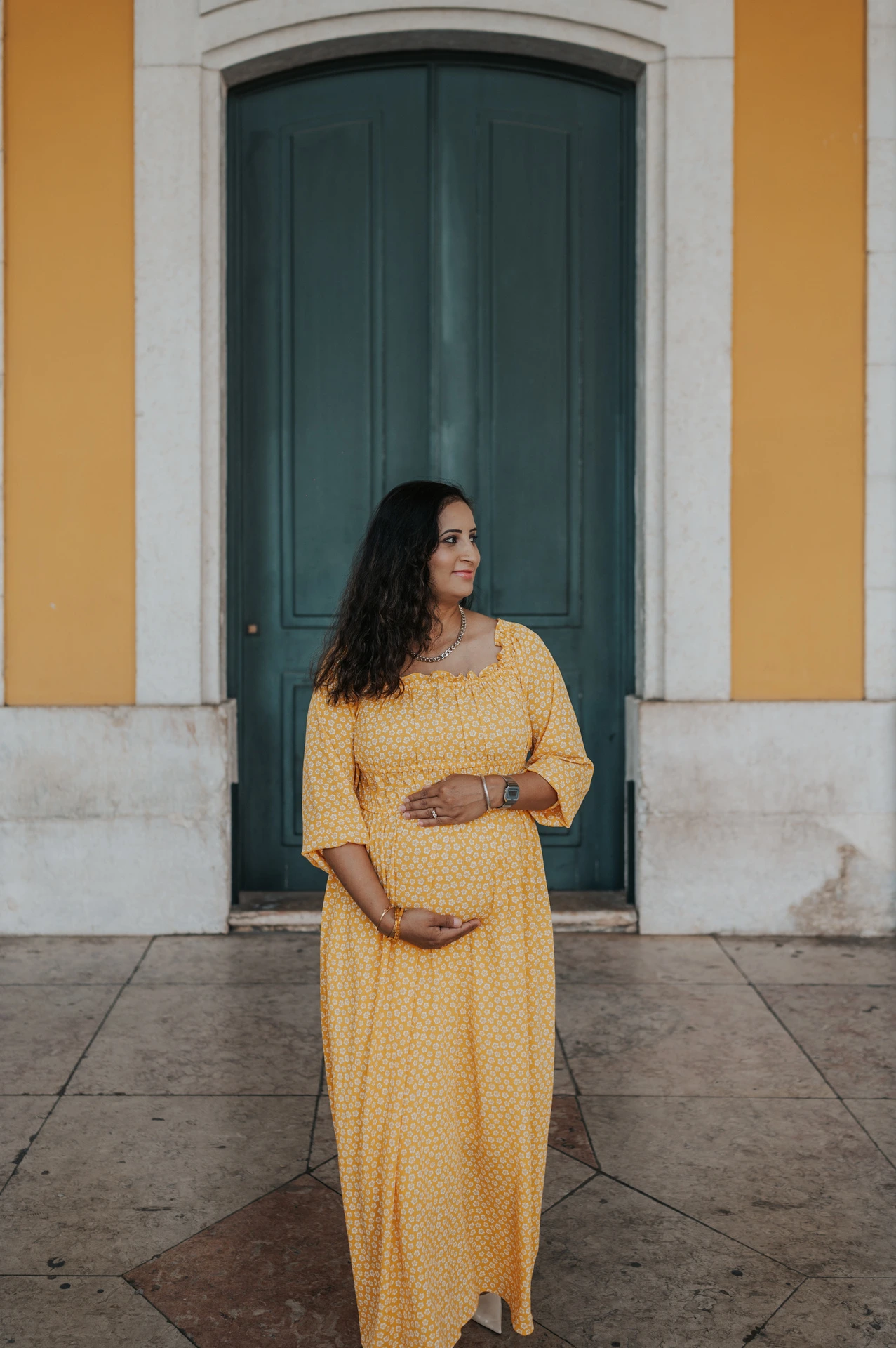 The expectant mother holding her belly in front of the tall green door at Praça do Comércio during their family and maternity photoshoot in Lisbon.
