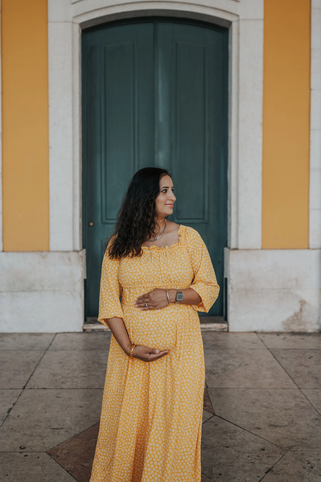 The expectant mother holding her belly in front of the tall green door at Praça do Comércio during their family and maternity photoshoot in Lisbon.