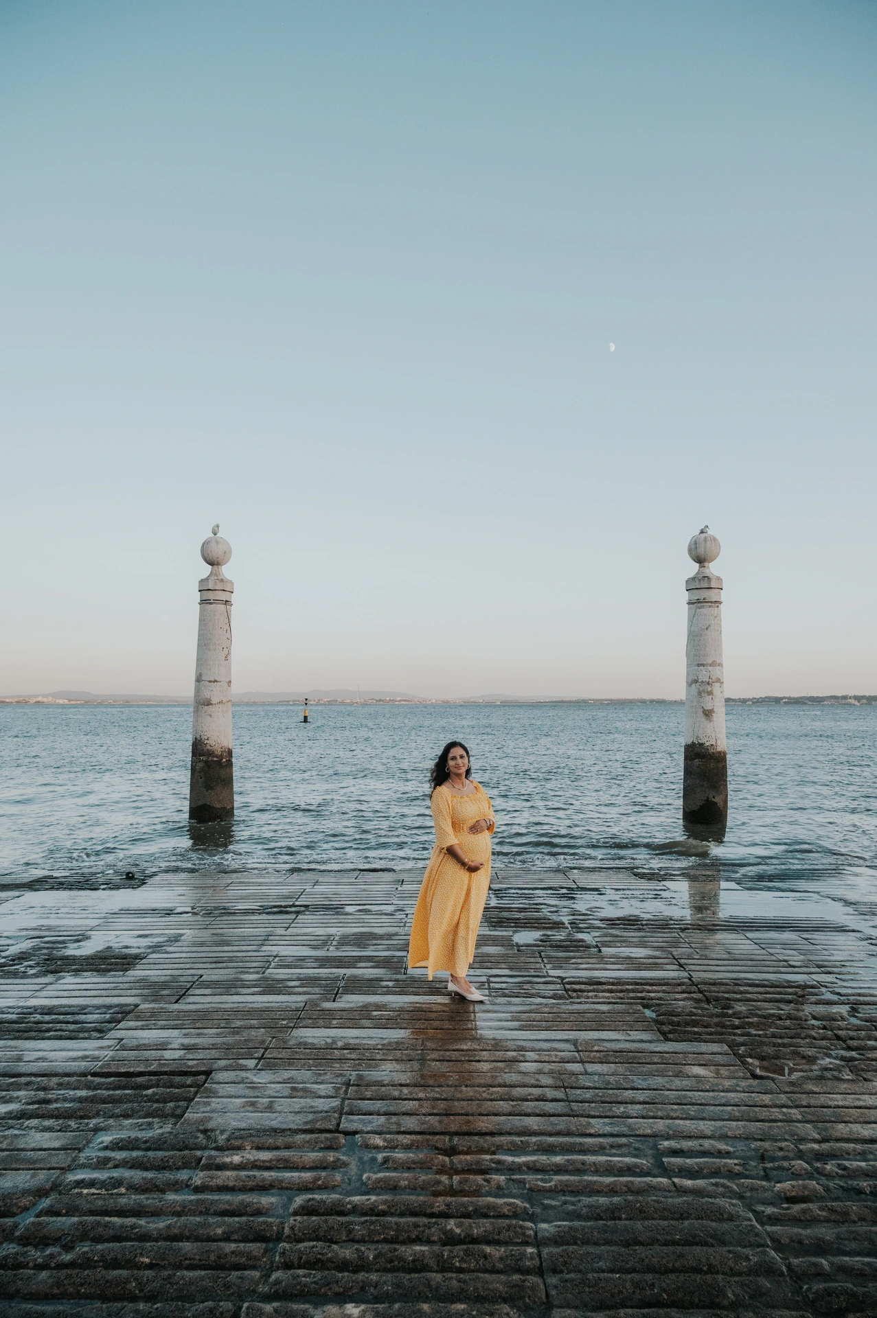 The expectant mother in a yellow dress holding her belly beside the Tagus River during their family and maternity photoshoot in Lisbon.