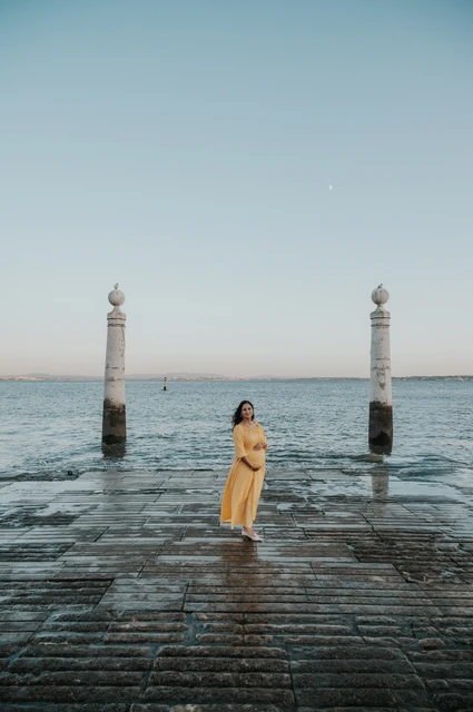 A maternity portrait beside the river | Lisbon family and maternity photoshoot The expectant mother in a yellow dress holding her belly beside the Tagus River during their family and maternity photoshoot in Lisbon.