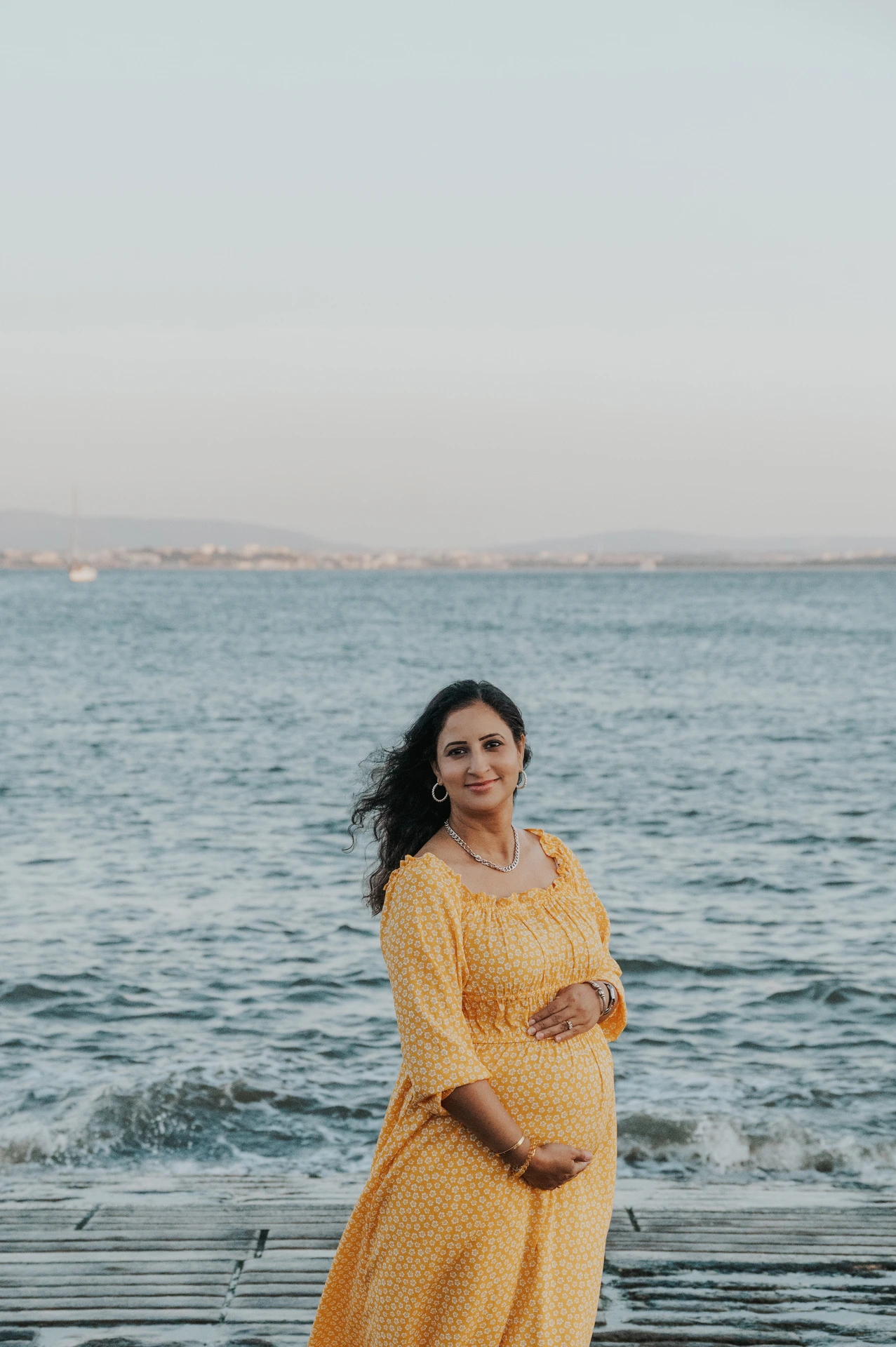 The expectant mother in a yellow dress holding her belly beside the Tagus River during their family and maternity photoshoot in Lisbon.