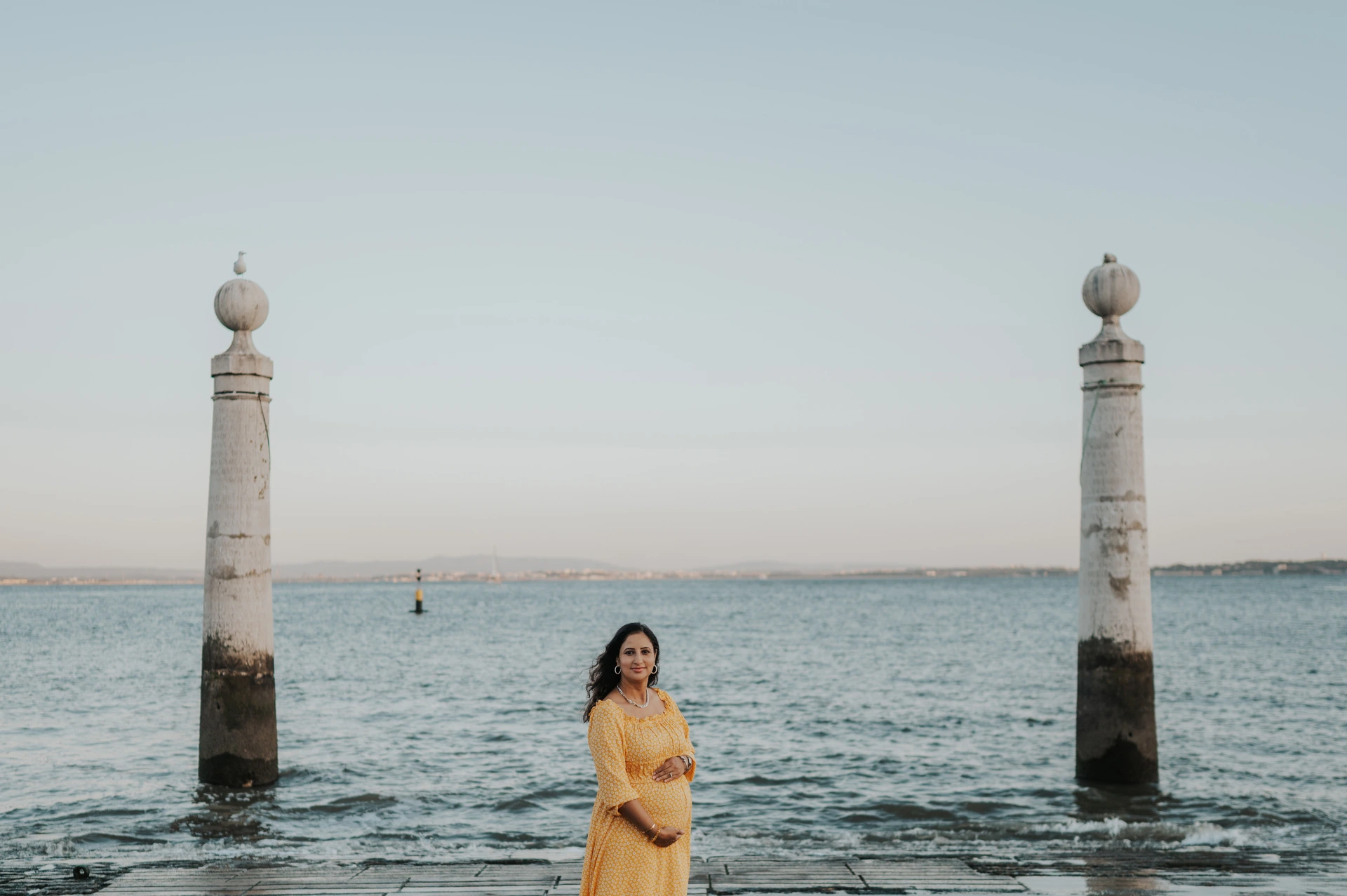 The expectant mother in a yellow dress holding her belly beside the Tagus River during their family and maternity photoshoot in Lisbon.
