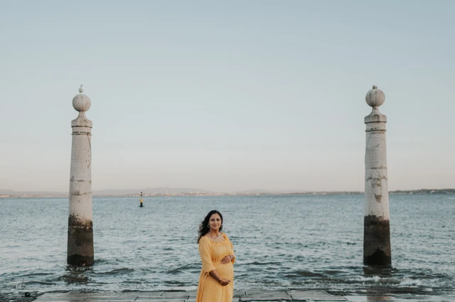 The expectant mother in a yellow dress holding her belly beside the Tagus River during their family and maternity photoshoot in Lisbon.