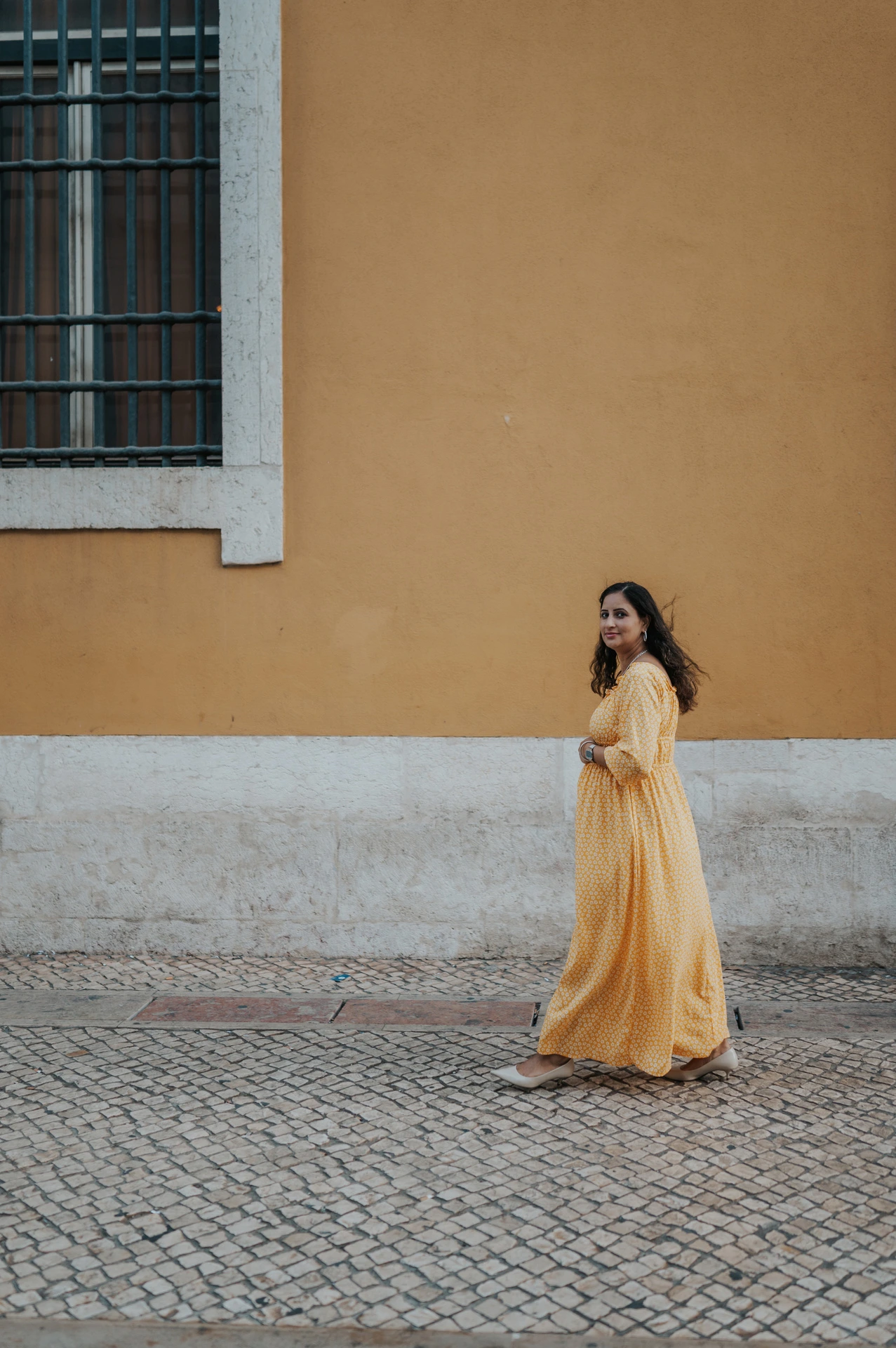 The expectant mother holding her belly beside the square’s yellow wall during their family and maternity photoshoot in Lisbon.