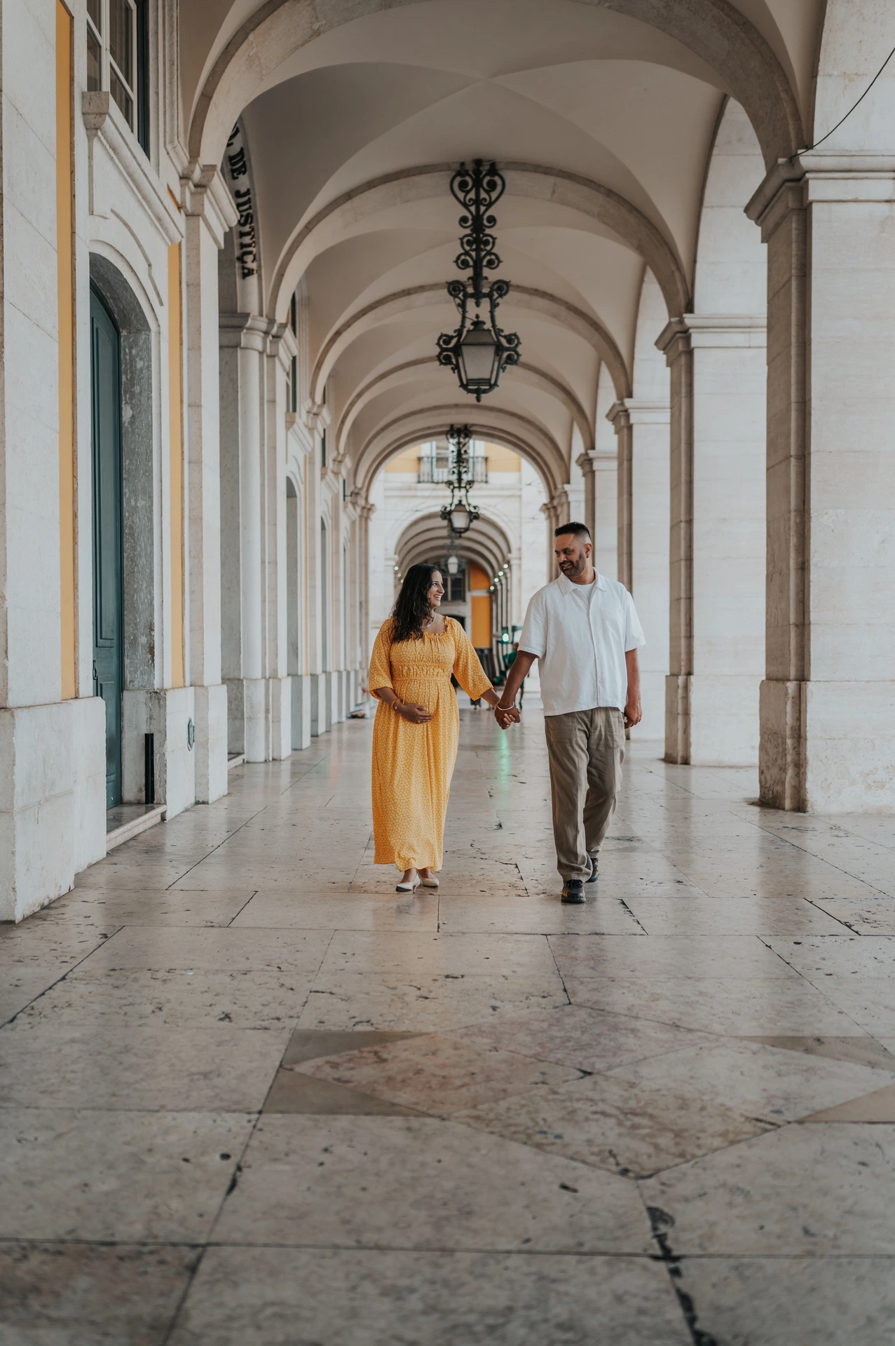 The expectant parents holding hands beneath the long stone arcade at Praça do Comércio during their family and maternity photoshoot in Lisbon.