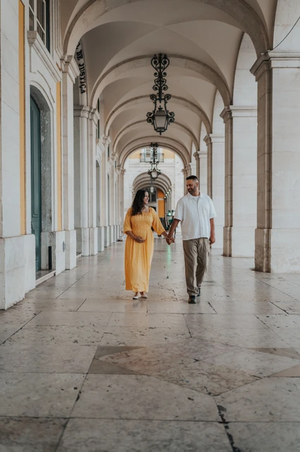 A walk beneath the square’s arches | Lisbon family and maternity photoshoot The expectant parents holding hands beneath the long stone arcade at Praça do Comércio during their family and maternity photoshoot in Lisbon.