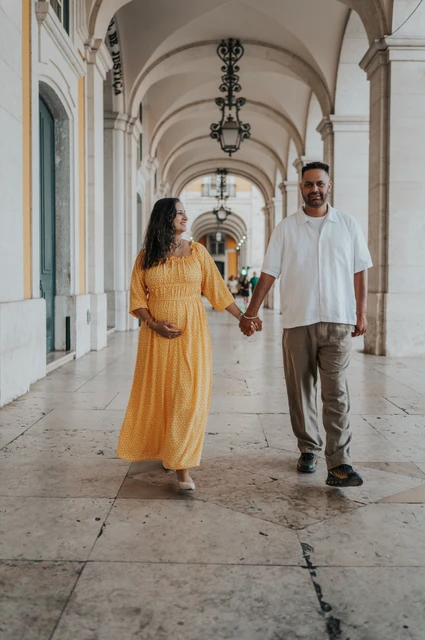 A walk beneath the square’s arches | Lisbon family and maternity photoshoot The expectant parents holding hands beneath the long stone arcade at Praça do Comércio during their family and maternity photoshoot in Lisbon.