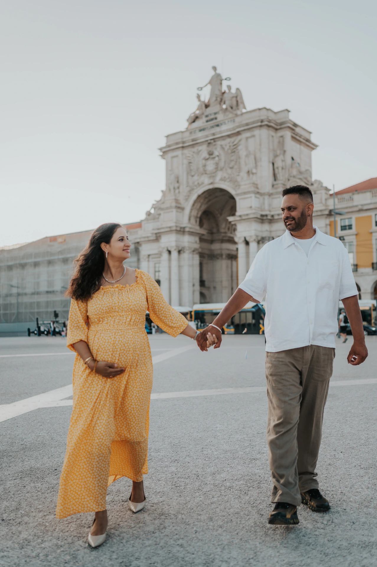 The expectant parents holding hands with Praça do Comércio’s arch behind them during their family and maternity photoshoot in Lisbon.