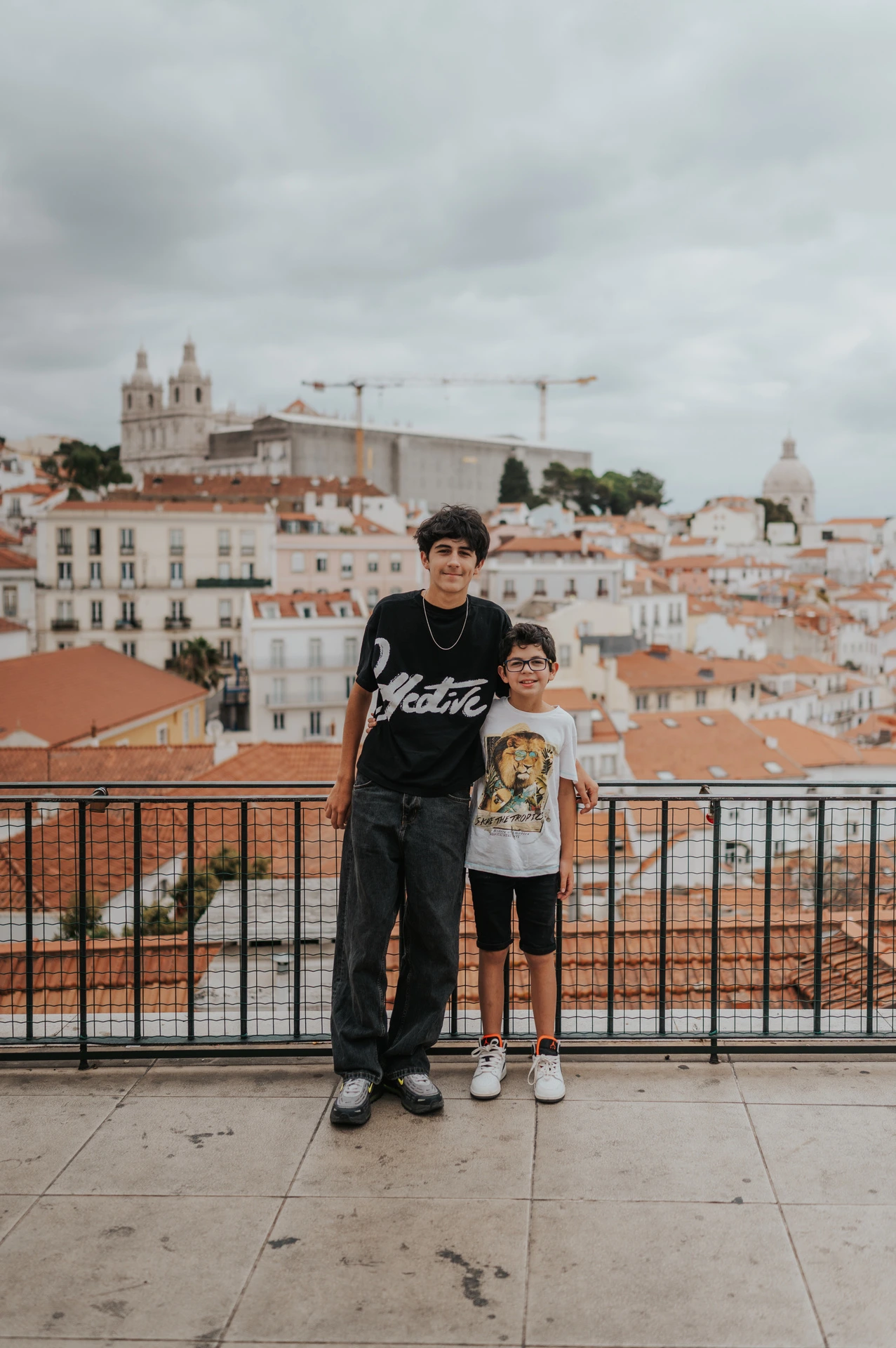 The two children standing together beside the viewpoint railing with Lisbon’s rooftops behind them during their family photoshoot in Lisbon.