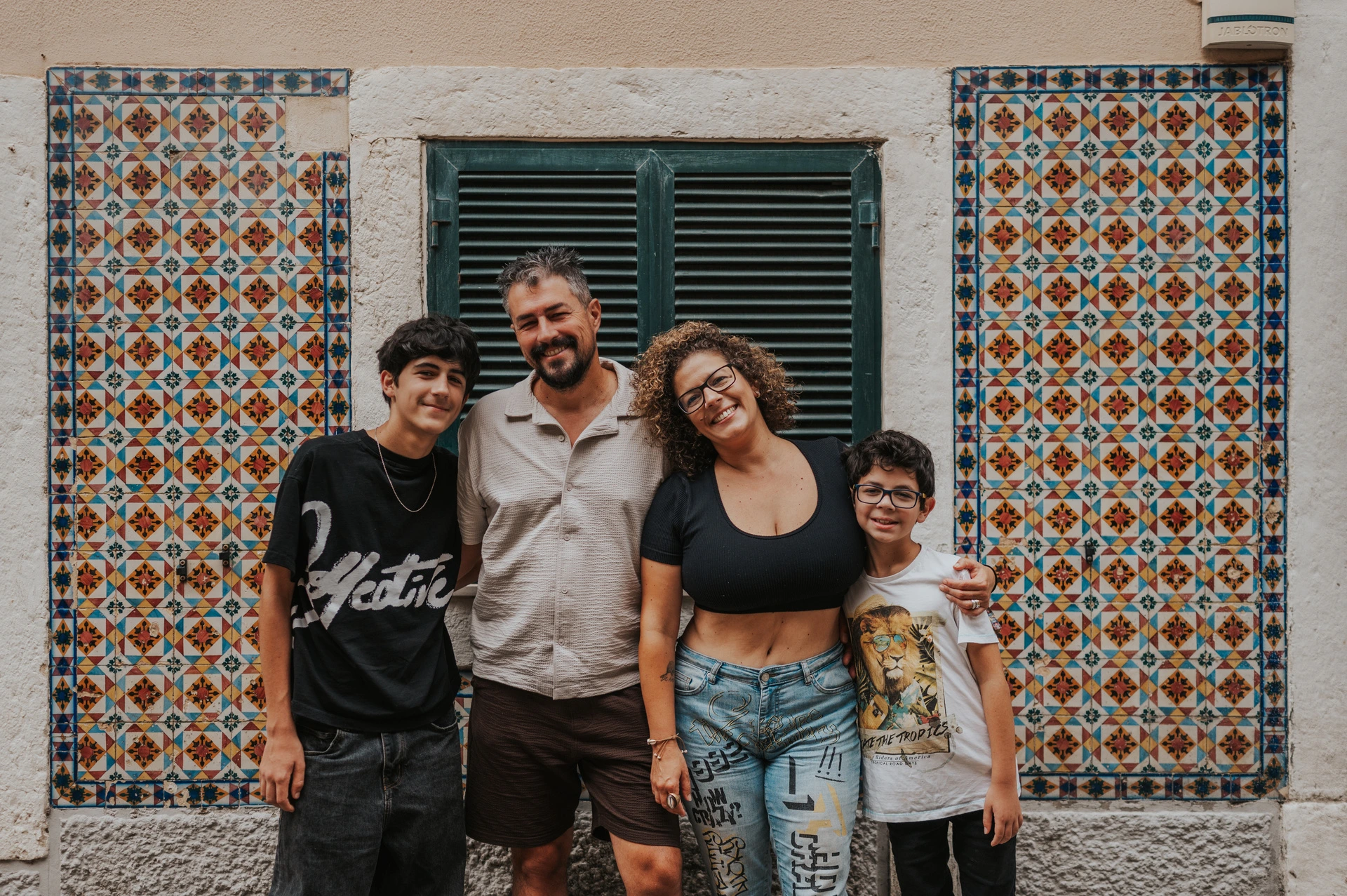 The family standing together against a colourful patterned tile wall and green shutters during their family photoshoot in Lisbon.