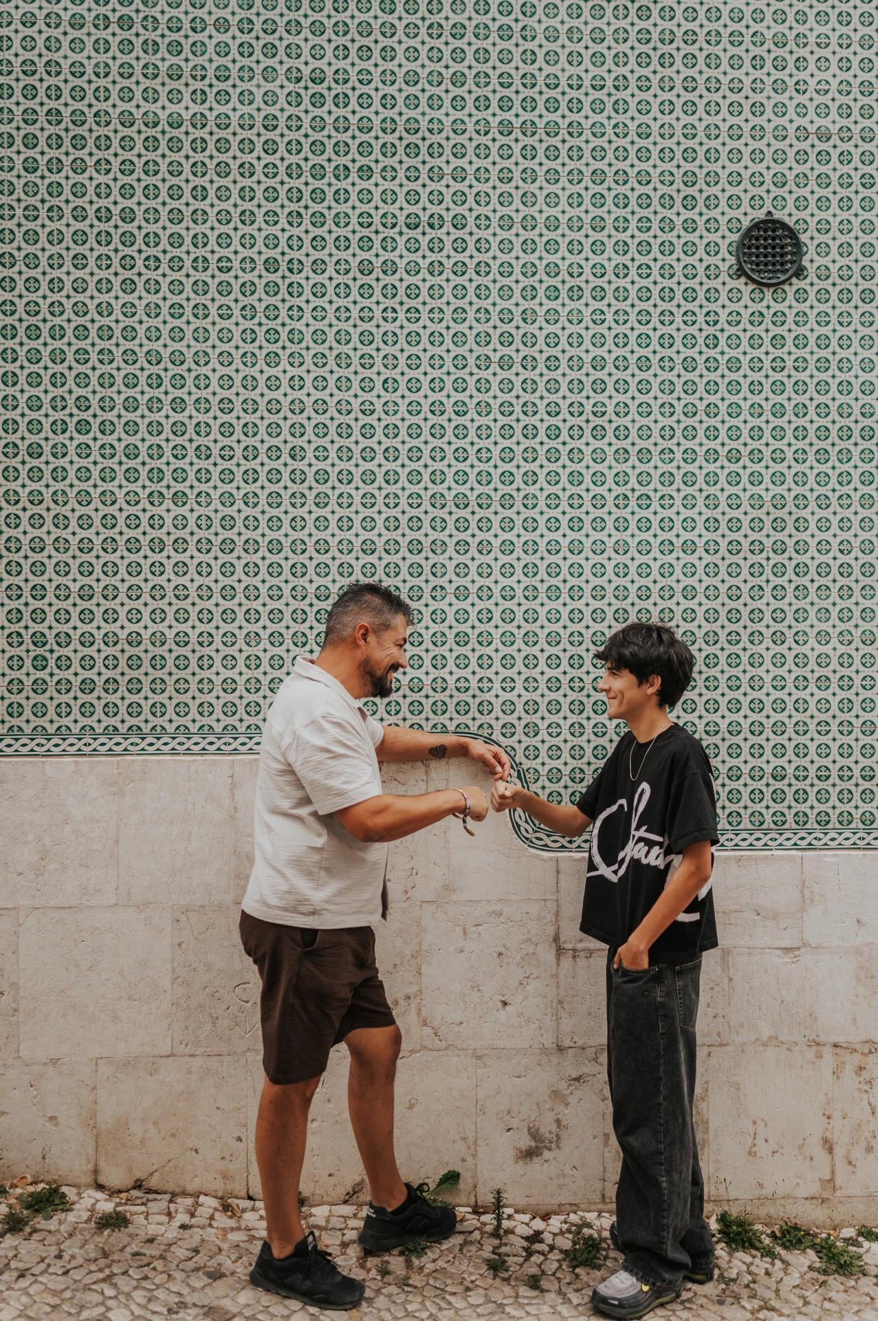A parent smiling with the older child beside a green patterned tile wall during their family photoshoot in Lisbon.