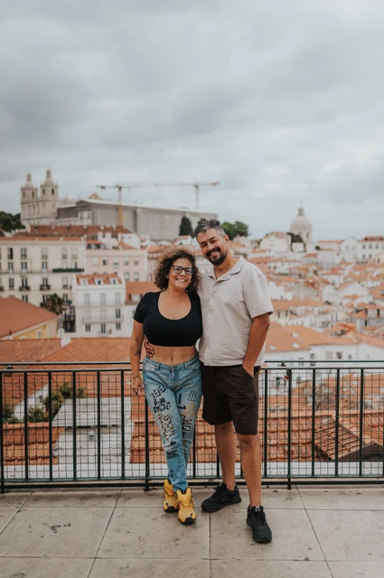 A portrait of the parents above the city | Lisbon family photoshoot The parents standing close together and smiling beside the viewpoint railing during their family photoshoot in Lisbon.