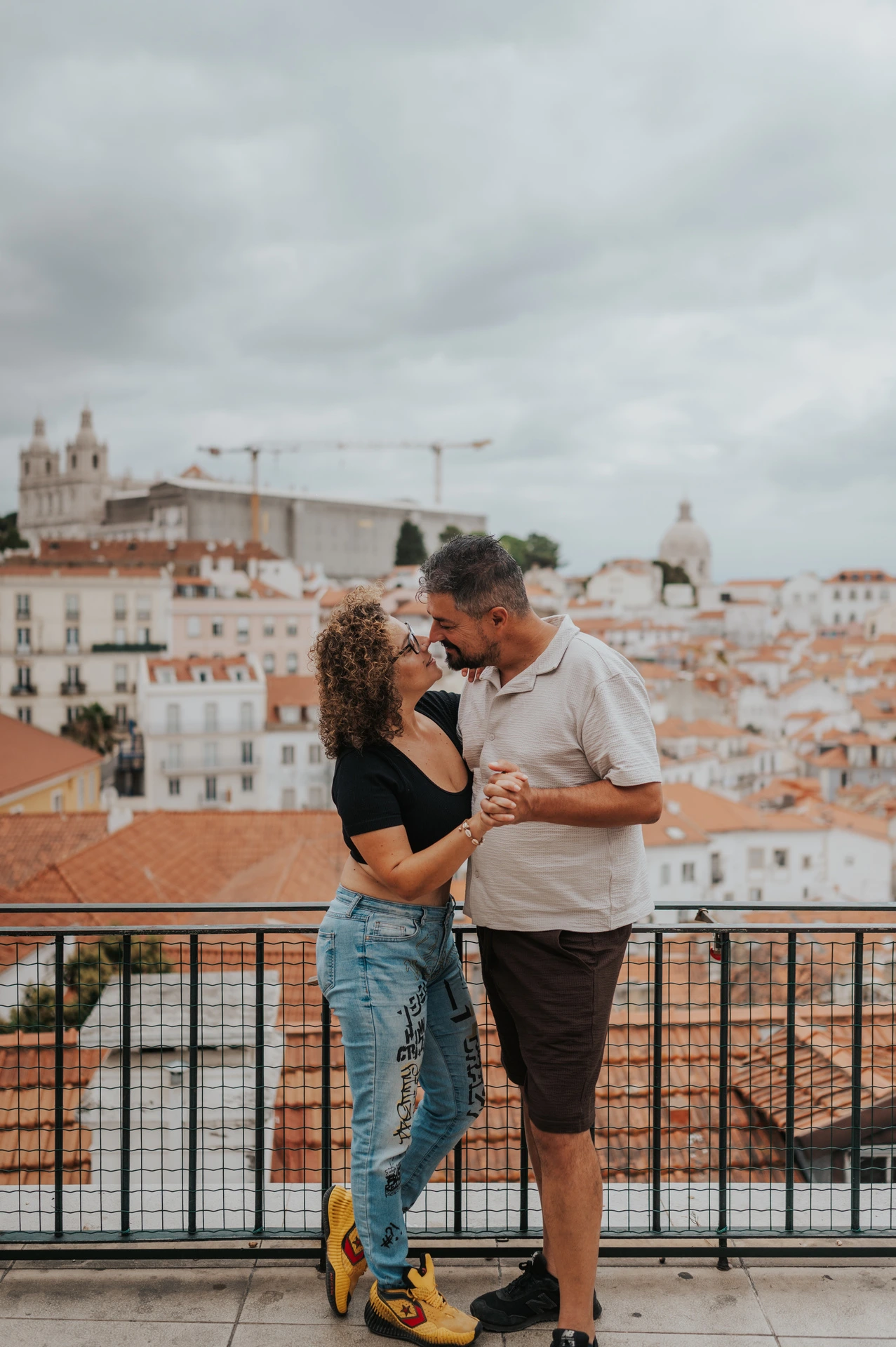 The parents standing close together and smiling beside the viewpoint railing during their family photoshoot in Lisbon.