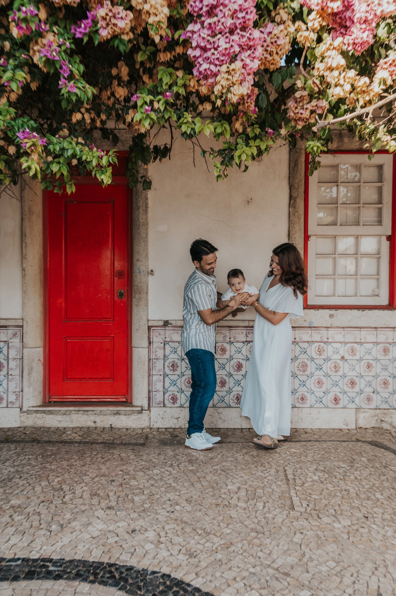 The parents holding their child up between them beneath the flowering branches at the red doorway during their family photoshoot in Lisbon.