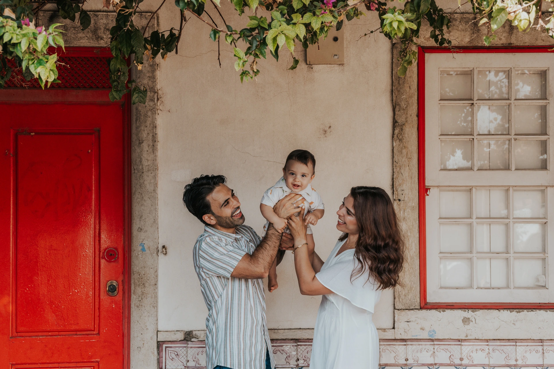 A smiling child beneath the flowers | Lisbon family photoshoot The parents holding their child up between them beneath the flowering branches at the red doorway during their family photoshoot in Lisbon.