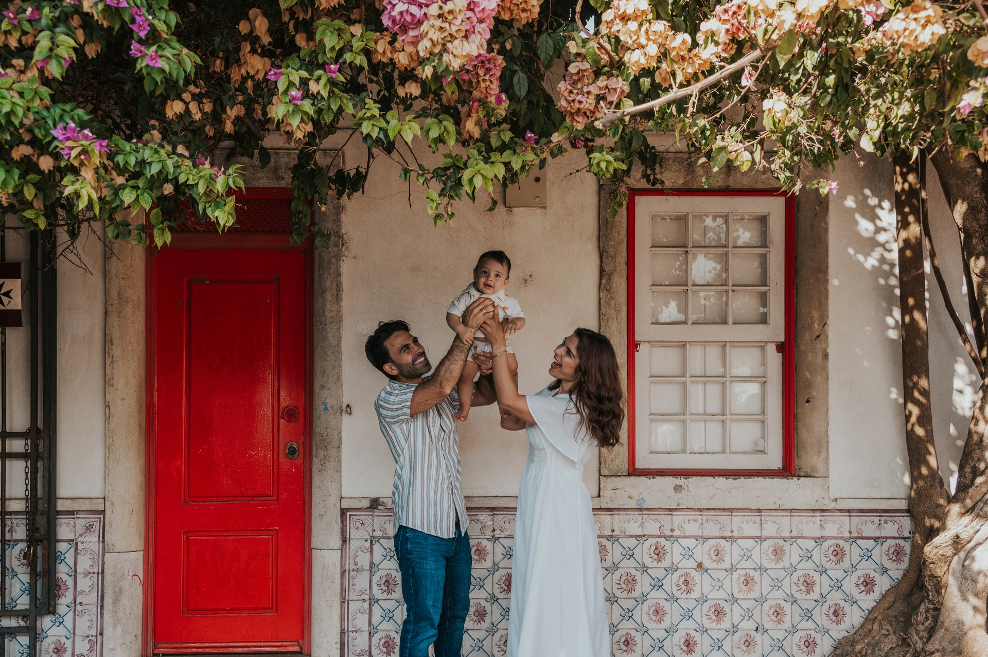The parents holding their child up between them beneath the flowering branches at the red doorway during their family photoshoot in Lisbon.