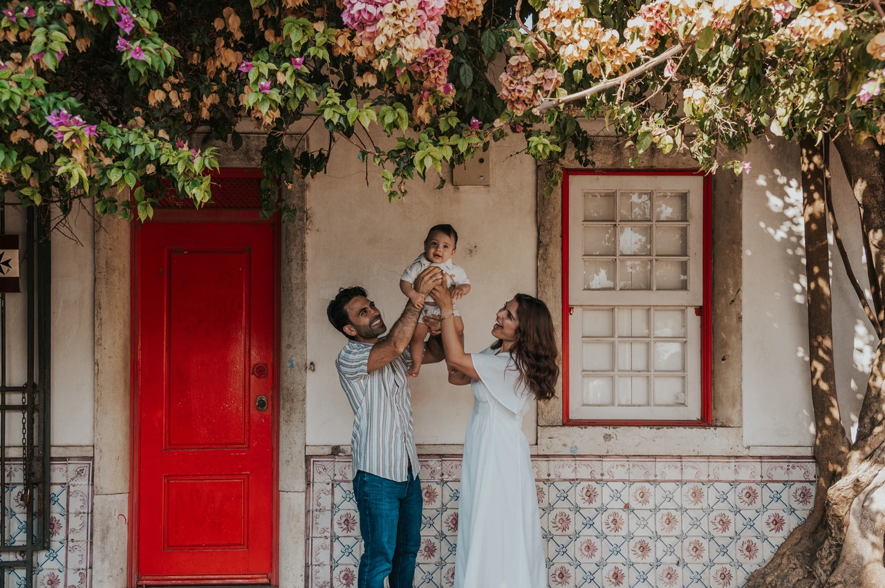 A smiling child beneath the flowers | Lisbon family photoshoot The parents holding their child up between them beneath the flowering branches at the red doorway during their family photoshoot in Lisbon.