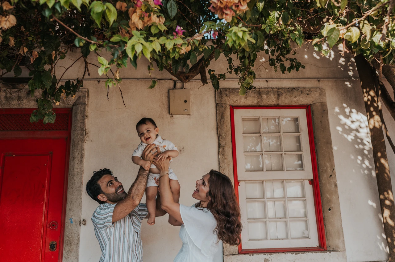 A smiling child beneath the flowers | Lisbon family photoshoot The parents holding their child up between them beneath the flowering branches at the red doorway during their family photoshoot in Lisbon.