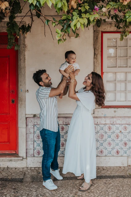 A smiling child beneath the flowers | Lisbon family photoshoot The parents holding their child up between them beneath the flowering branches at the red doorway during their family photoshoot in Lisbon.
