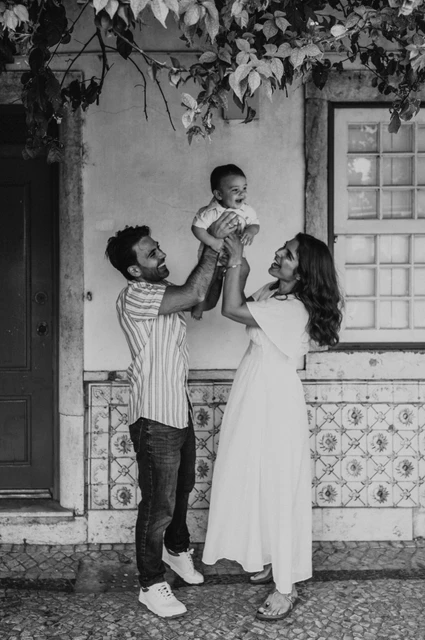 A smiling child beneath the flowers in black and white | Lisbon family photoshoot Black-and-white photograph: the parents holding their child up between them beneath the flowering branches at the red doorway during their family photoshoot in Lisbon.