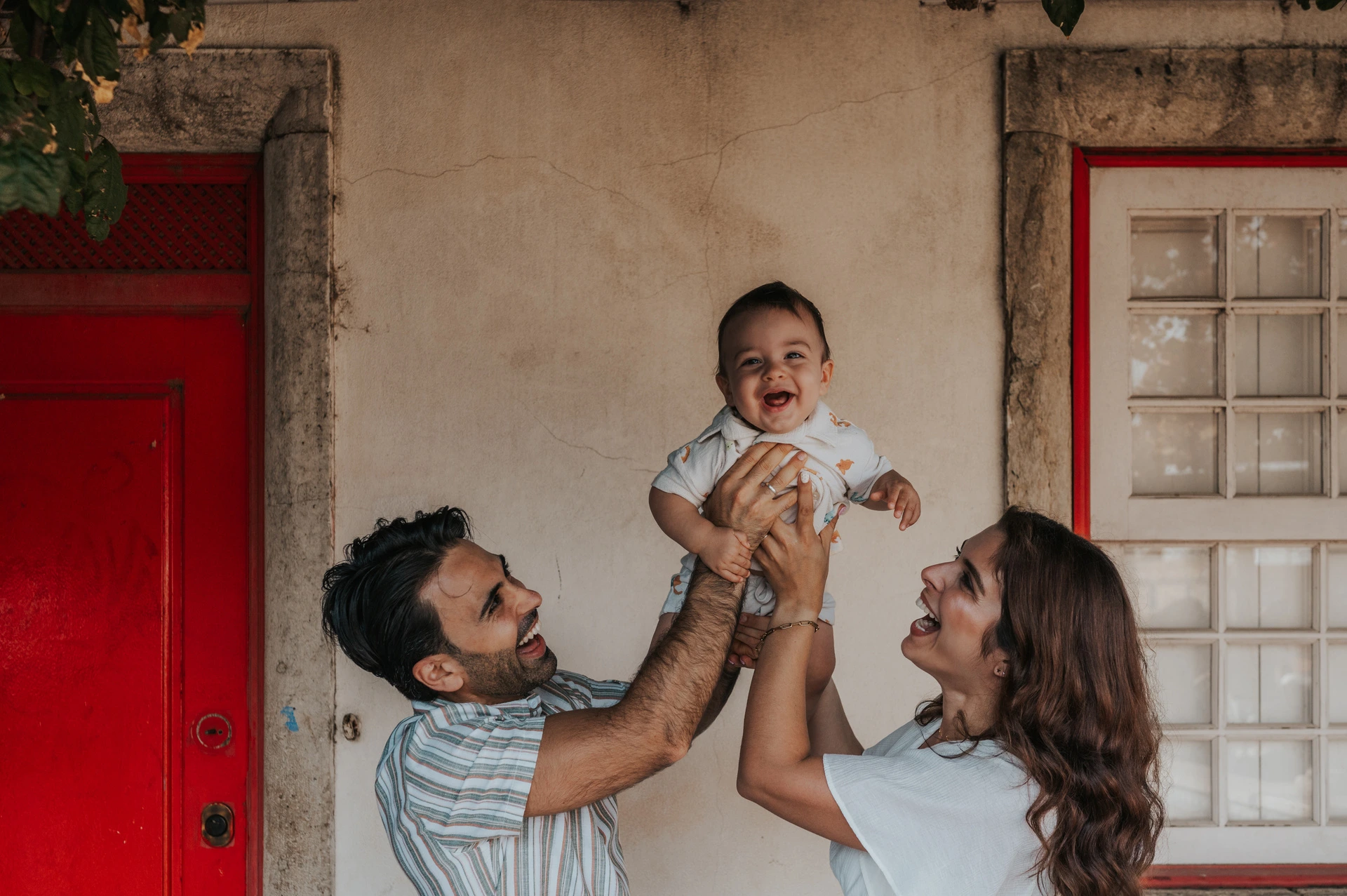 The parents holding their child up between them beneath the flowering branches at the red doorway during their family photoshoot in Lisbon.