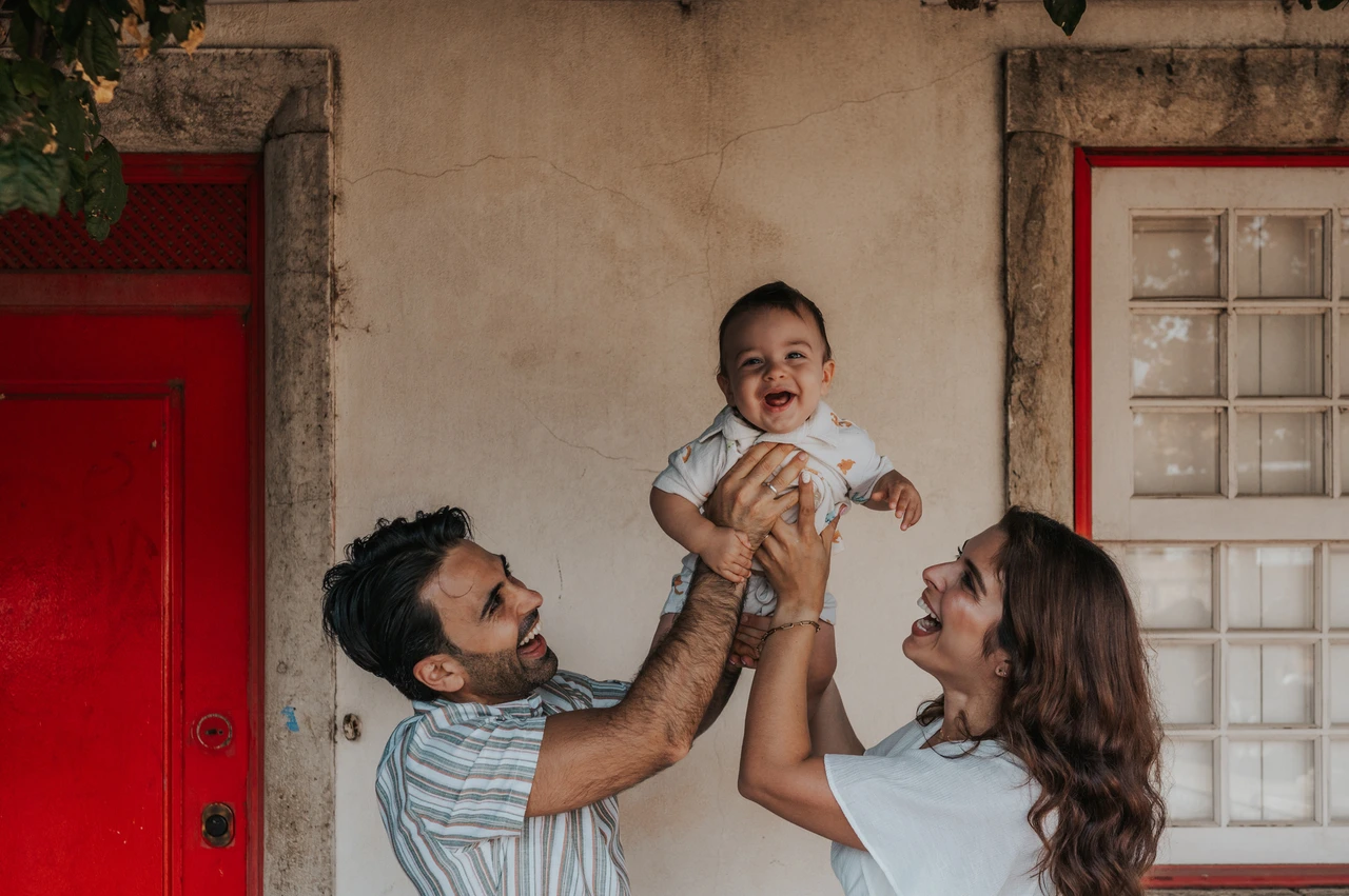 A smiling child beneath the flowers | Lisbon family photoshoot The parents holding their child up between them beneath the flowering branches at the red doorway during their family photoshoot in Lisbon.