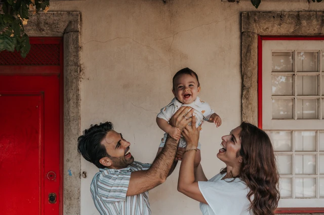 The parents holding their child up between them beneath the flowering branches at the red doorway during their family photoshoot in Lisbon.