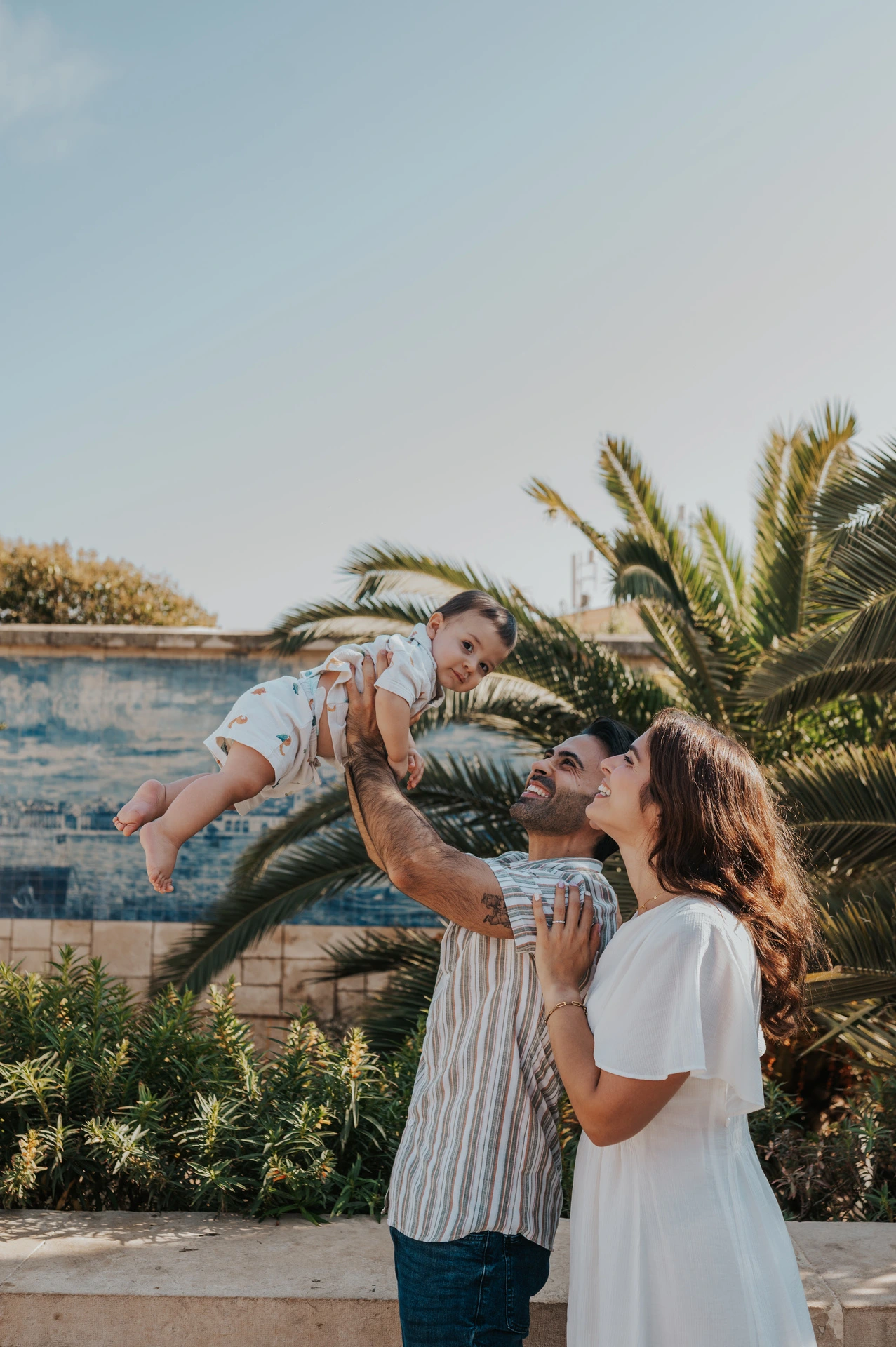 The parents lifting their smiling child between them beside palms and blue tiles during their family photoshoot in Lisbon.